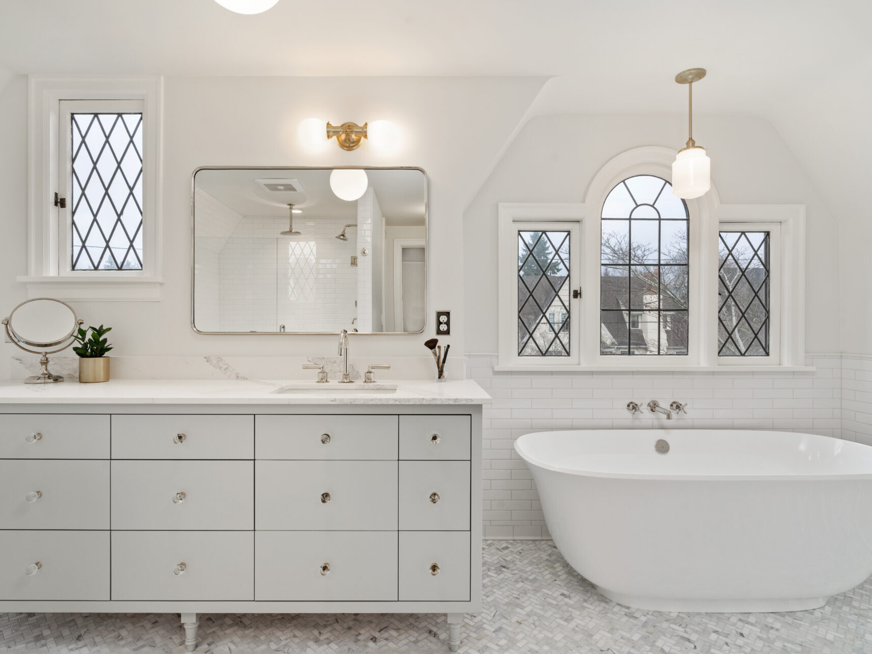 A bright, elegant bathroom in Portland, Oregon features a large, freestanding white bathtub by the windows. A white vanity with multiple drawers, two sinks, and a potted plant graces the space alongside a round magnifying mirror. Mosaic tiles beautifully cover the floor.