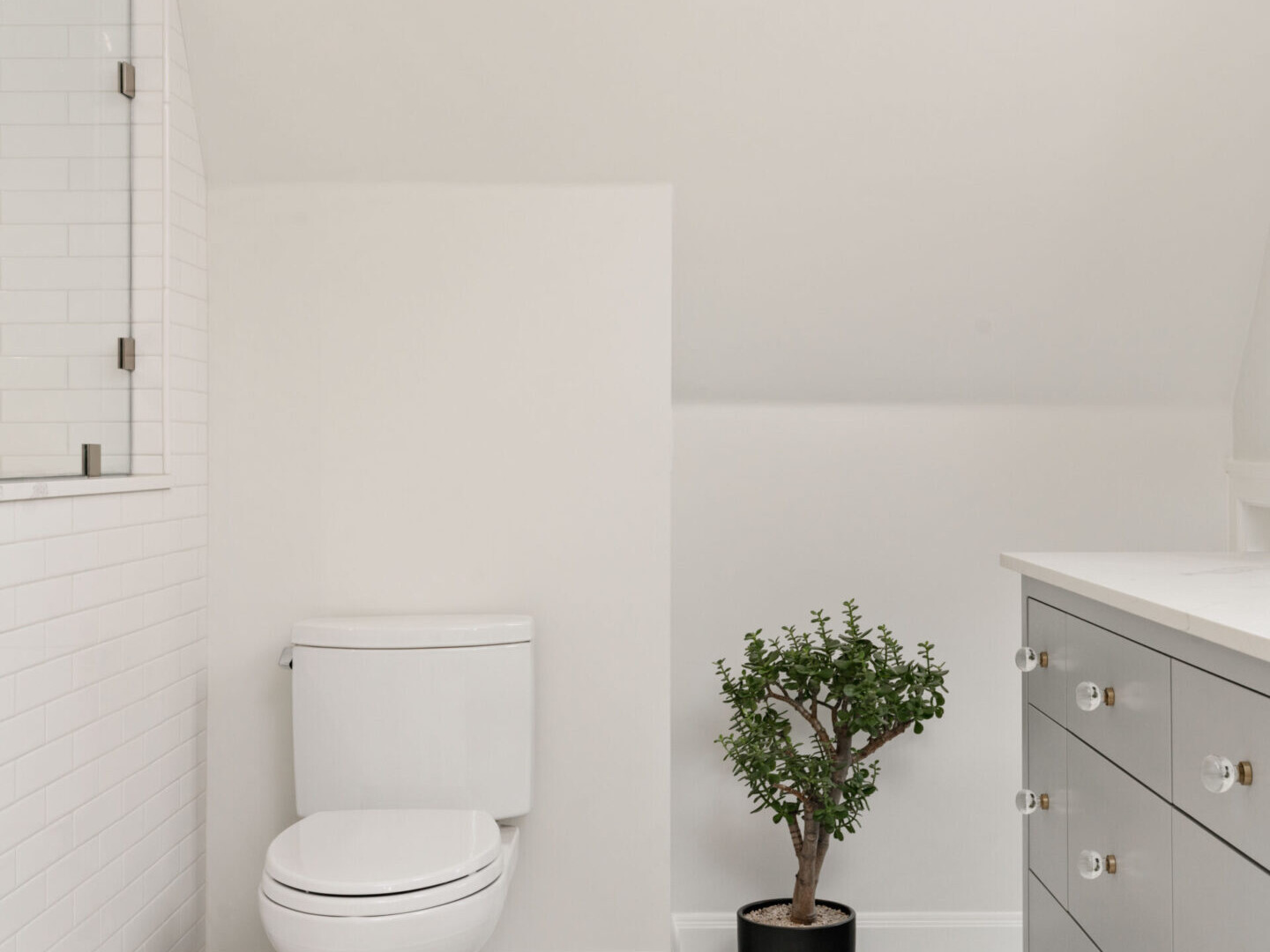 A minimalist bathroom in Portland, Oregon features a white toilet beside a small potted plant. The space boasts a light-colored tiled floor and a vanity with gray cabinets and round knobs, complemented by light walls and a sleek glass shower enclosure.