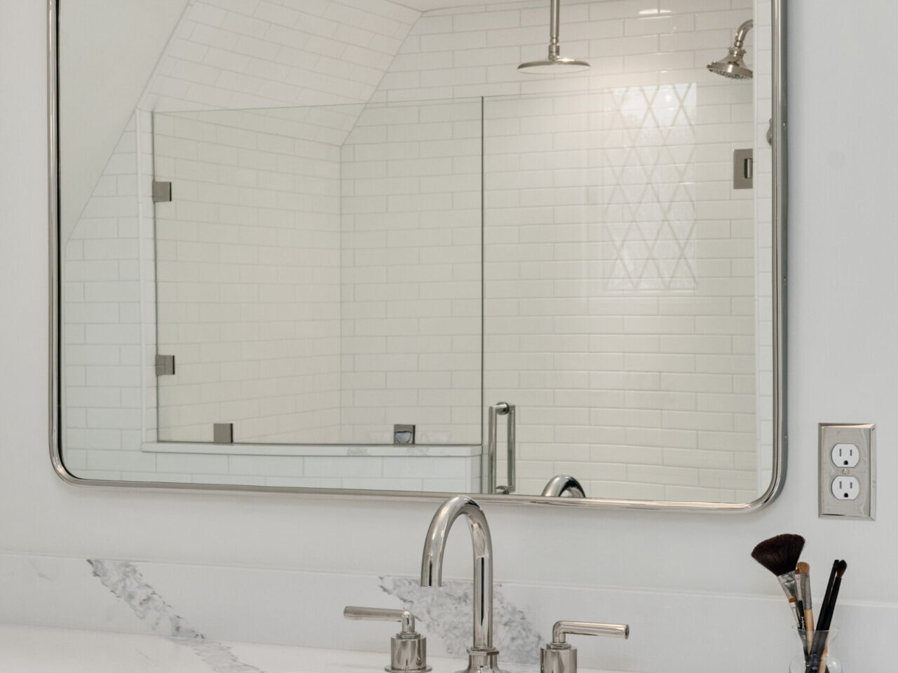 A modern bathroom in Portland, Oregon, showcases a large rectangular mirror above a sleek countertop with a marble finish. The sink has a silver faucet, and the light fixture above the mirror has two round bulbs. White subway tiles line the walls.