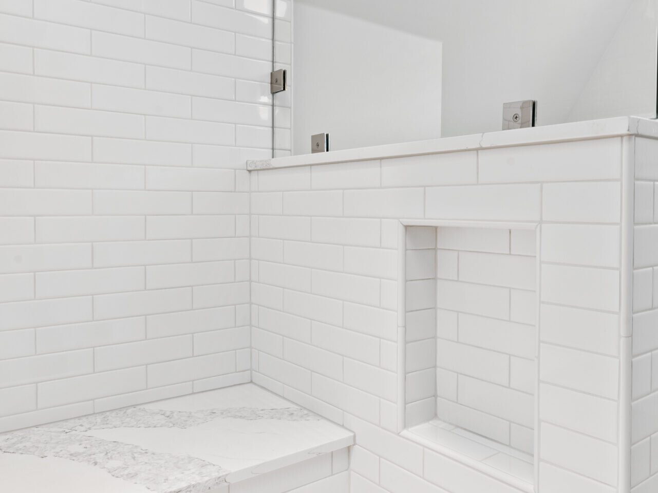 A bright, white-tiled shower in Portland, Oregon, featuring a built-in bench and recessed shelf. The tiles are arranged in a horizontal brick pattern, and the space is beautifully illuminated by natural light. The floor showcases an intricate mosaic tile design.