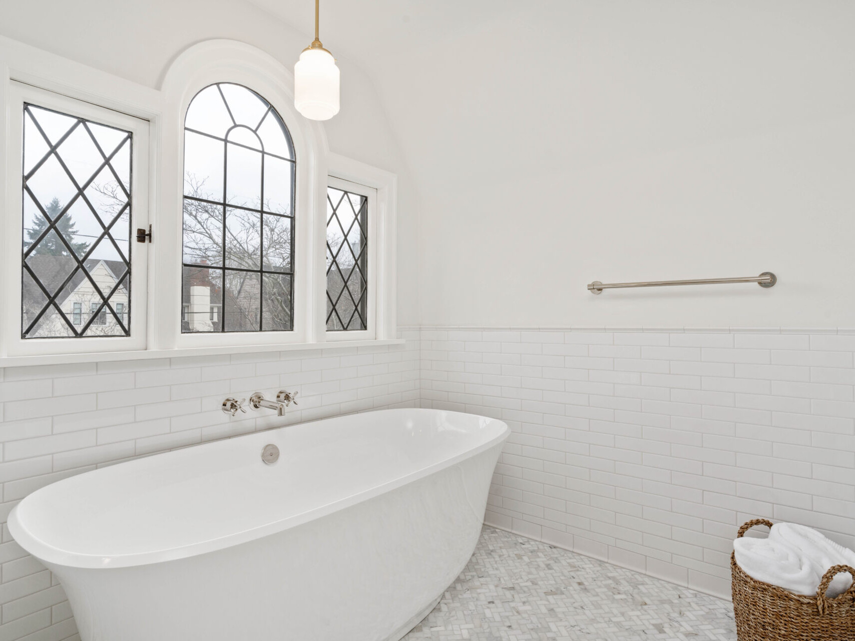 A bright bathroom in Portland, Oregon features a white freestanding bathtub, white subway tiles on the walls, and a large arched window with diamond-patterned panes. A woven basket with towels sits beside the tub, and a towel bar is mounted on the wall.