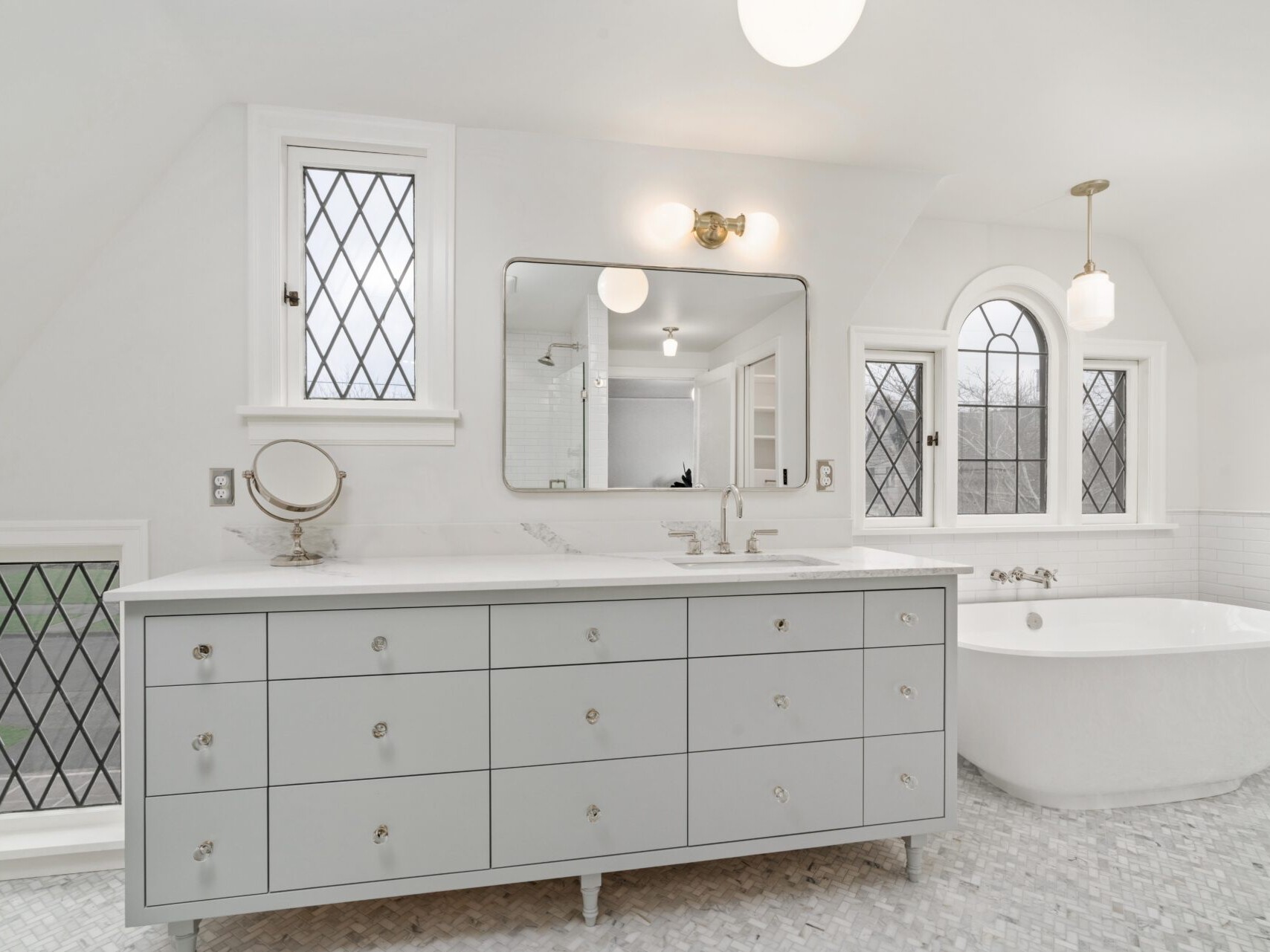 A bright bathroom in Portland, Oregon, features a white vanity with multiple drawers, a large mirror, and a freestanding white bathtub. Diamond-patterned windows accent the space, along with round lights and a convenient makeup mirror on the counter.