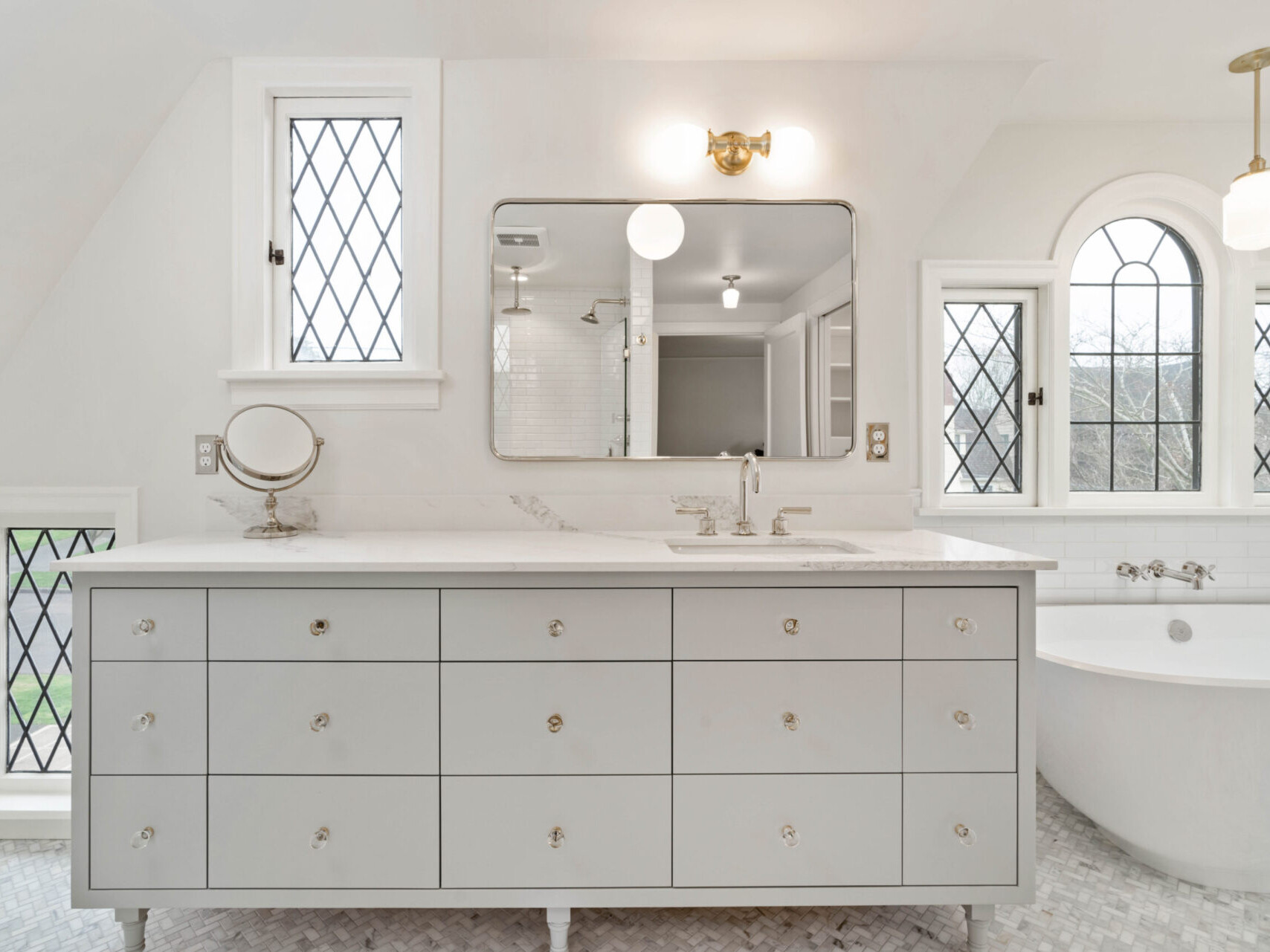 A spacious, elegant bathroom in Portland, Oregon features a large vanity with drawers, a mirror, wall sconces, and a freestanding bathtub. Three diamond-patterned windows provide natural light and a scenic view, enhancing the serene atmosphere.