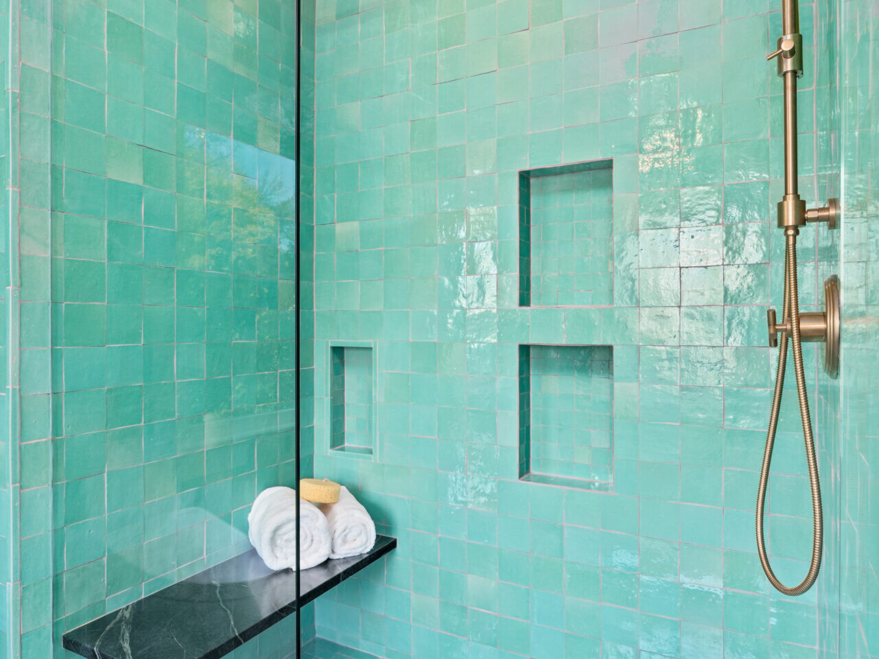 A modern shower in Portland, Oregon features turquoise tiles, a rain shower head, and a built-in glass panel. Recessed shelves line the wall, and a black marble bench is topped with neatly folded white towels.