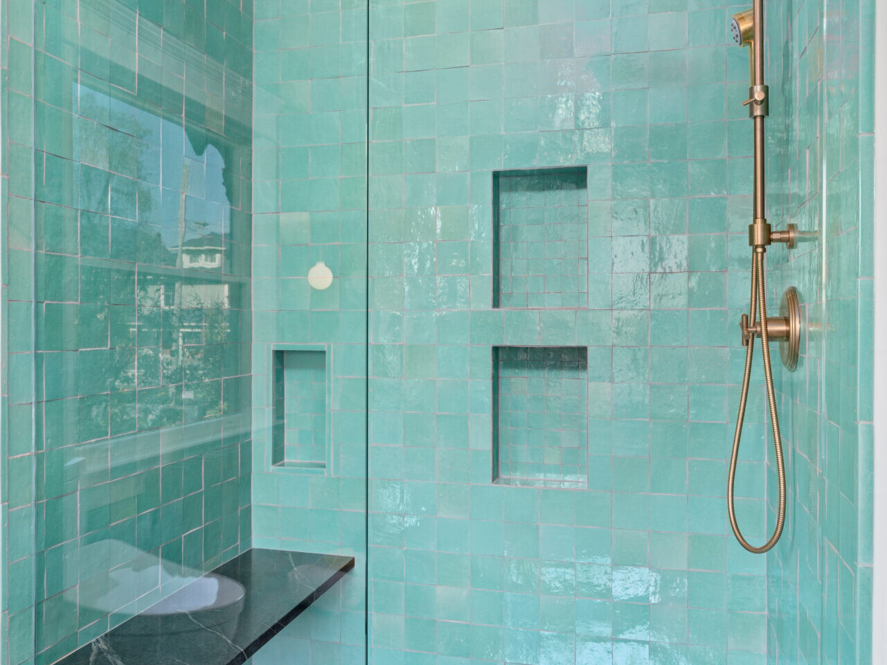 A modern shower in Portland, Oregon, with aqua tiles and a glass door. The wall features two built-in shelves and a bronze rain shower head. There's a black bench on the left side.