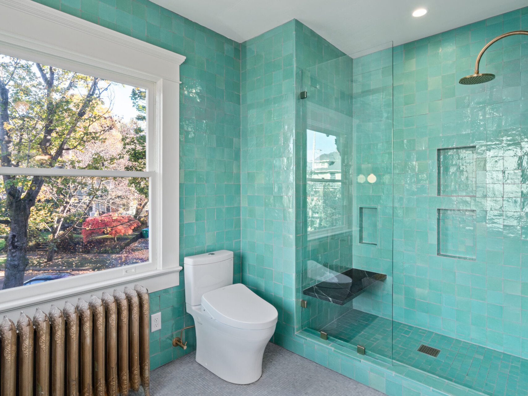 A bathroom in Portland, Oregon, boasts turquoise tiled walls and a glass-enclosed shower. A white toilet sits by a window with a view of trees outside. A bronze radiator and shower fixture provide an elegant contrast.