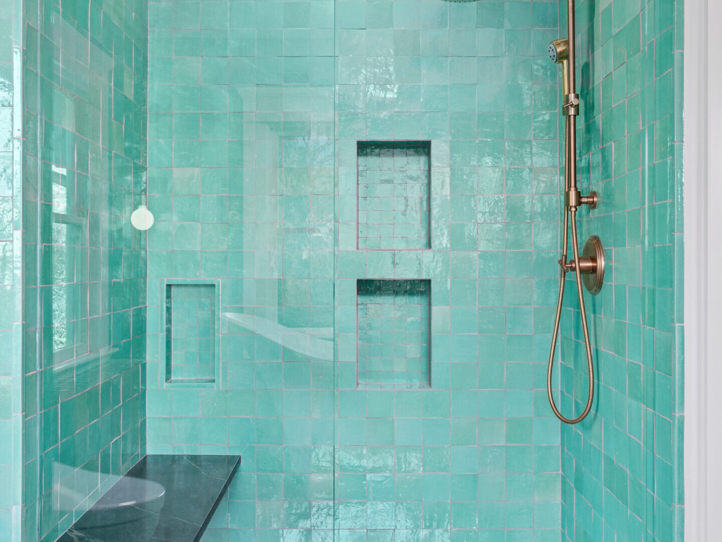 A modern shower in Portland, Oregon features teal tiles, a glass door, and a wall-mounted bronze rain showerhead. It includes two recessed shelves for storage and a built-in marble bench on the left side. The well-lit space is enhanced by sophisticated overhead lighting.