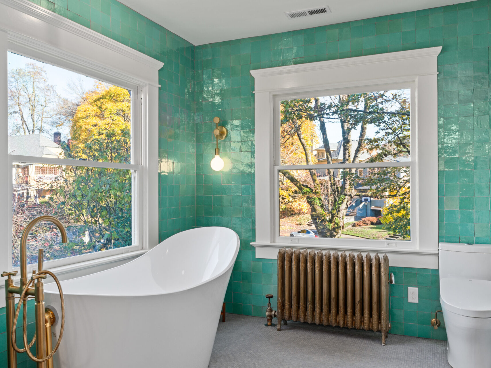 A bathroom in Portland, Oregon features teal tiled walls, a white freestanding bathtub with a gold faucet, and a large window overlooking trees. A vintage radiator sits below the window, paired with a spherical wall light to the left, complementing the serene atmosphere.
