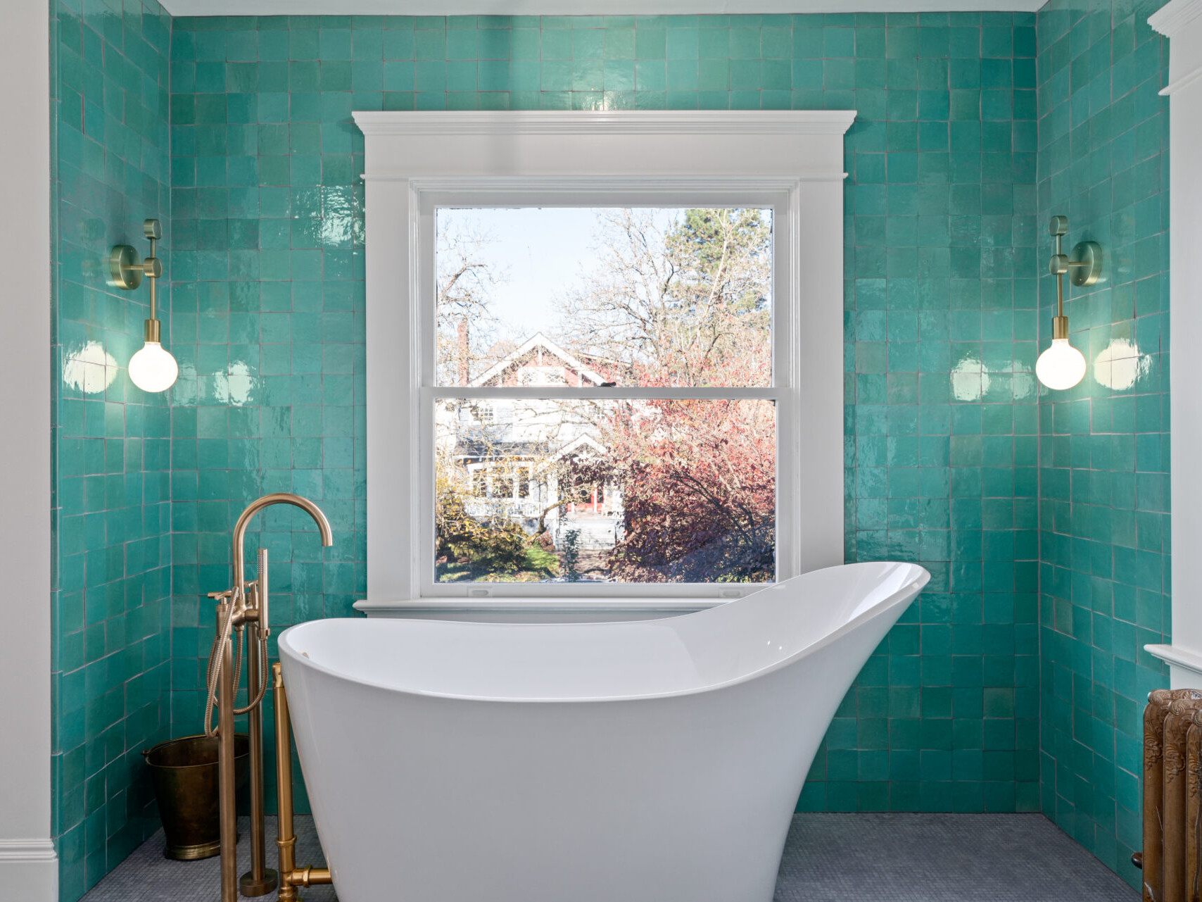 A modern bathroom in Portland, Oregon features a white freestanding bathtub in front of a window, with vibrant teal tiles on the walls. Two wall-mounted lights illuminate the space, and trees are visible outside through the window.