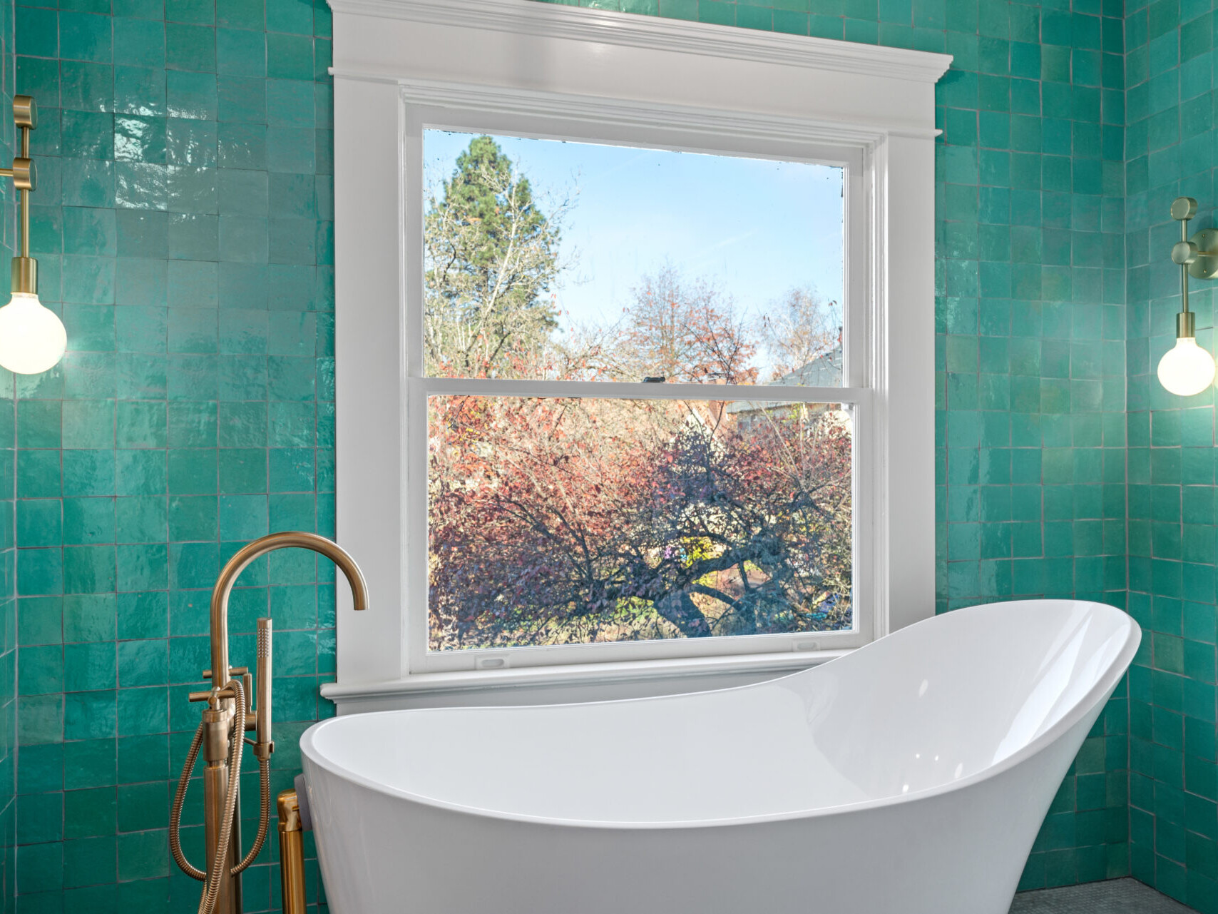 A modern bathroom with turquoise tiles, reminiscent of Portland, Oregon's eclectic style, features a white freestanding bathtub. Two brass faucets and matching wall sconces adorn either side. A window reveals a view of trees with autumn foliage.