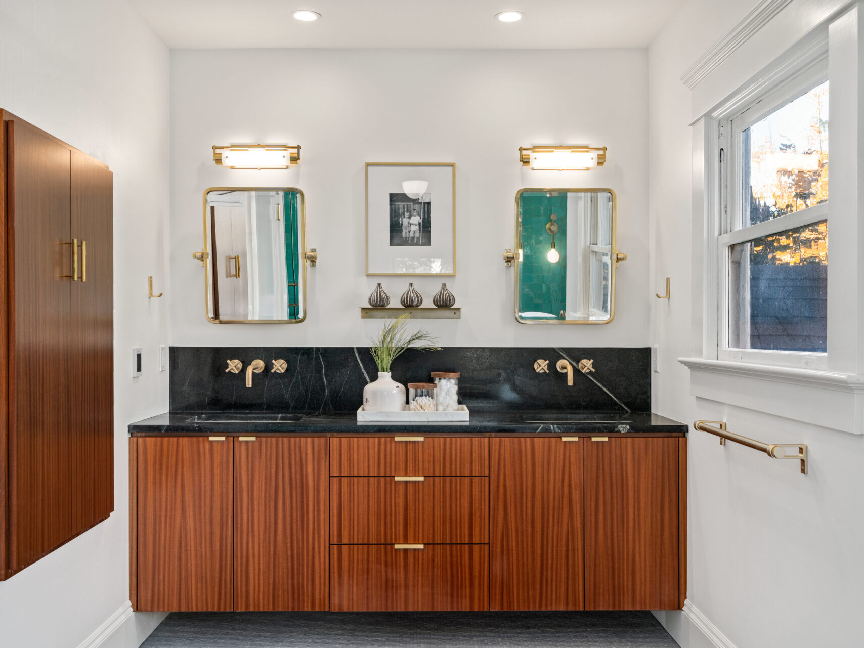 A modern bathroom in Portland, Oregon features a wooden double vanity with black stone countertop. Two mirrors are above the sinks, framed by wall sconces. A window on the right casts natural light, while a small art piece and decor sit elegantly between the mirrors.
