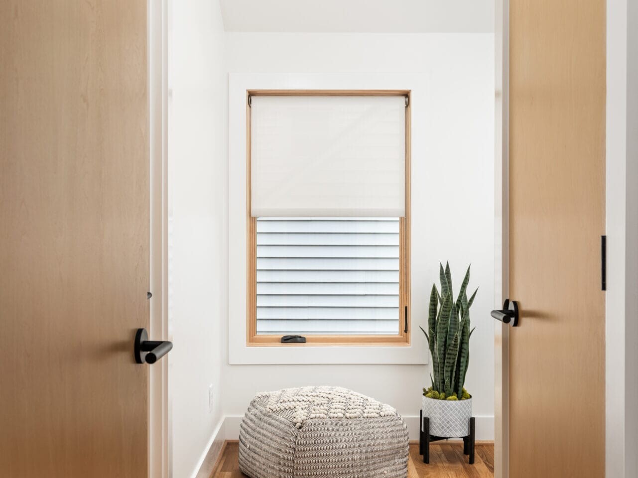 A minimalist room in Portland, Oregon, features wooden doors and flooring. There's a large window with a blind, a textured pouf, and a potted plant on the floor. The ceiling boasts a skylight, allowing natural light to fill the space.
