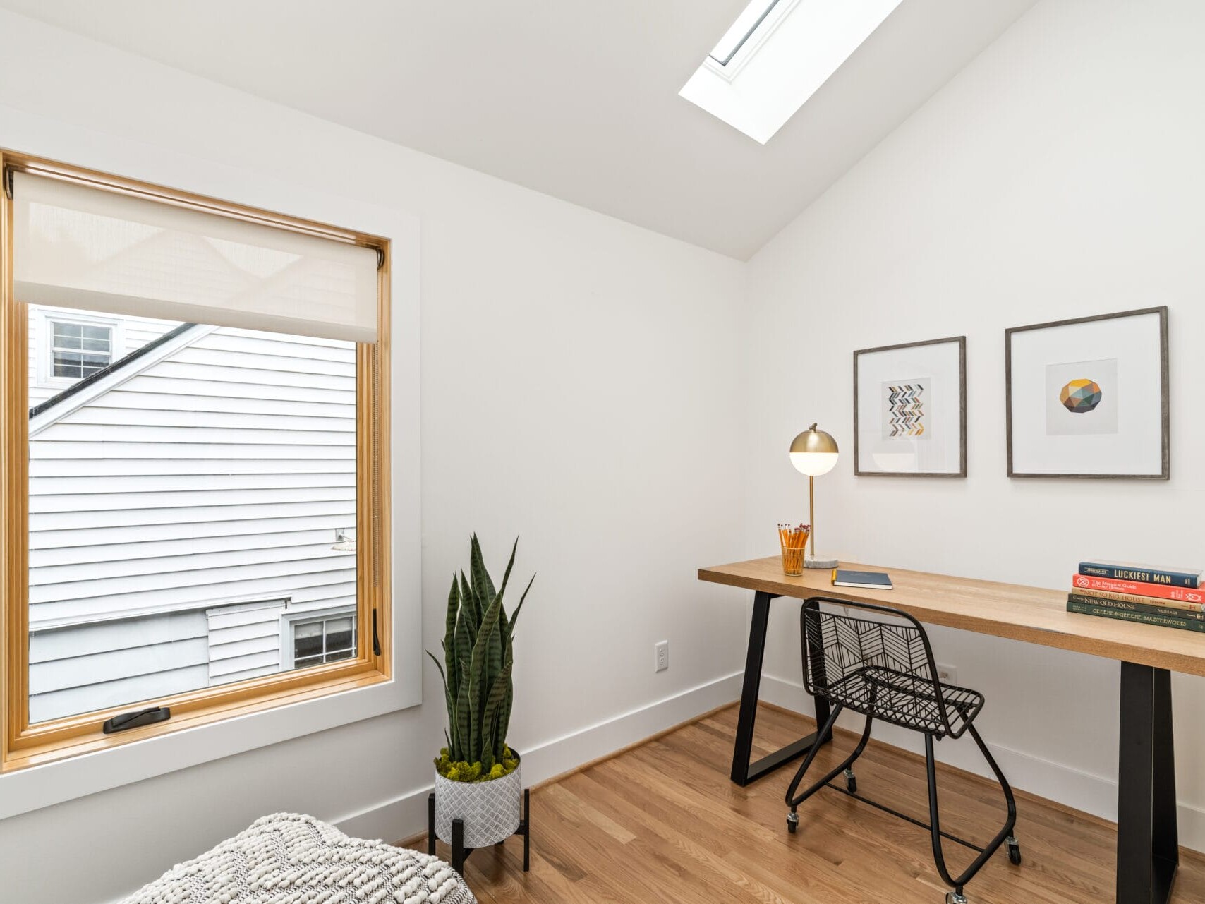 A minimalist Portland, Oregon home office features a wooden desk and black chair. A table lamp, books, and pencils adorn the desk. Two framed art pieces hang on the white wall. A plant sits beside a window, with natural light streaming in through a skylight.
