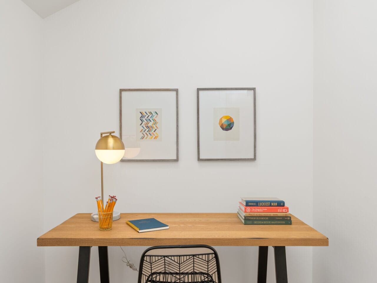 A small home office in Portland, Oregon features a wooden desk with a black chair. The desk holds a lamp, pencils, a notebook, and a stack of books. Two framed artworks adorn the white wall. Both a skylight and wall-mounted light offer ample illumination.