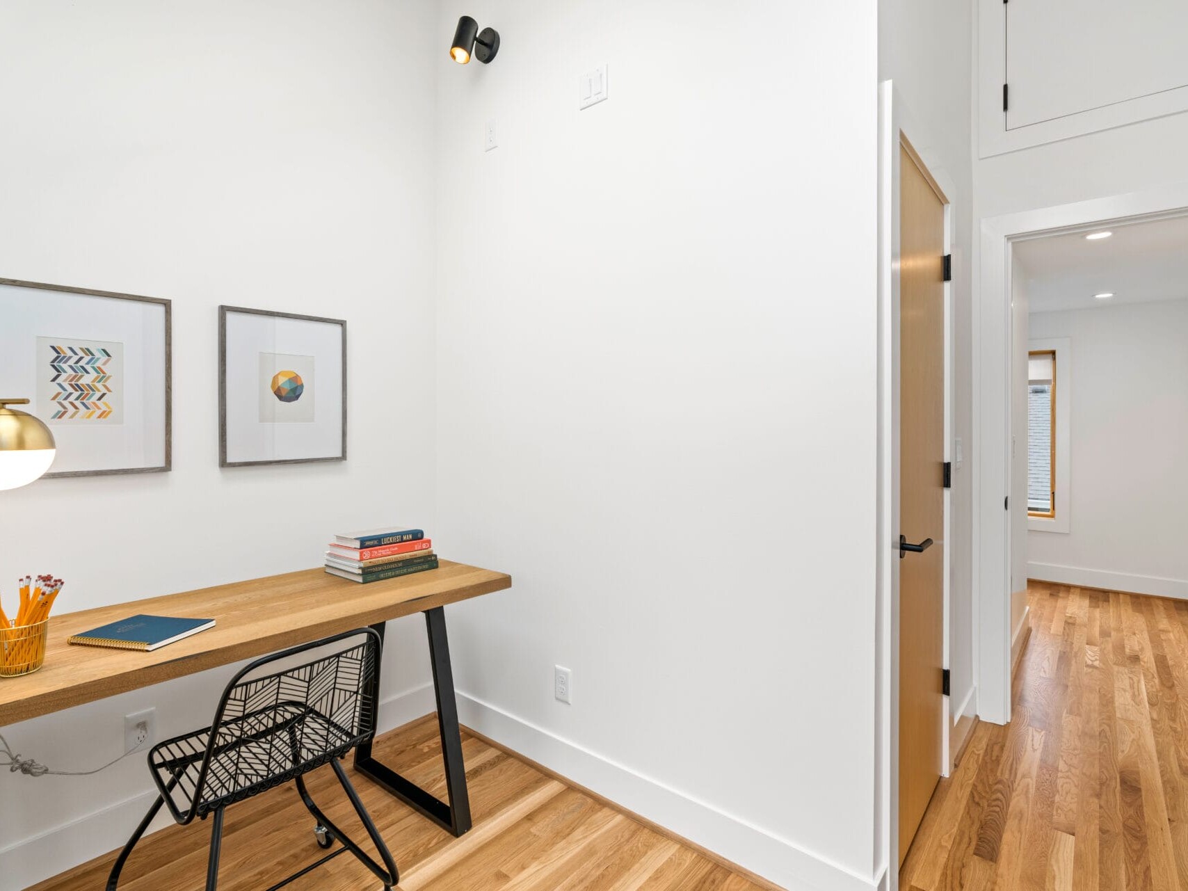 A modern, minimalist study space reminiscent of Portland, Oregon, featuring a wooden desk with a black wire chair. The desk holds books, a notebook, and a lamp. Two framed artworks hang on the white wall. A hallway leads to another room with wooden floors.