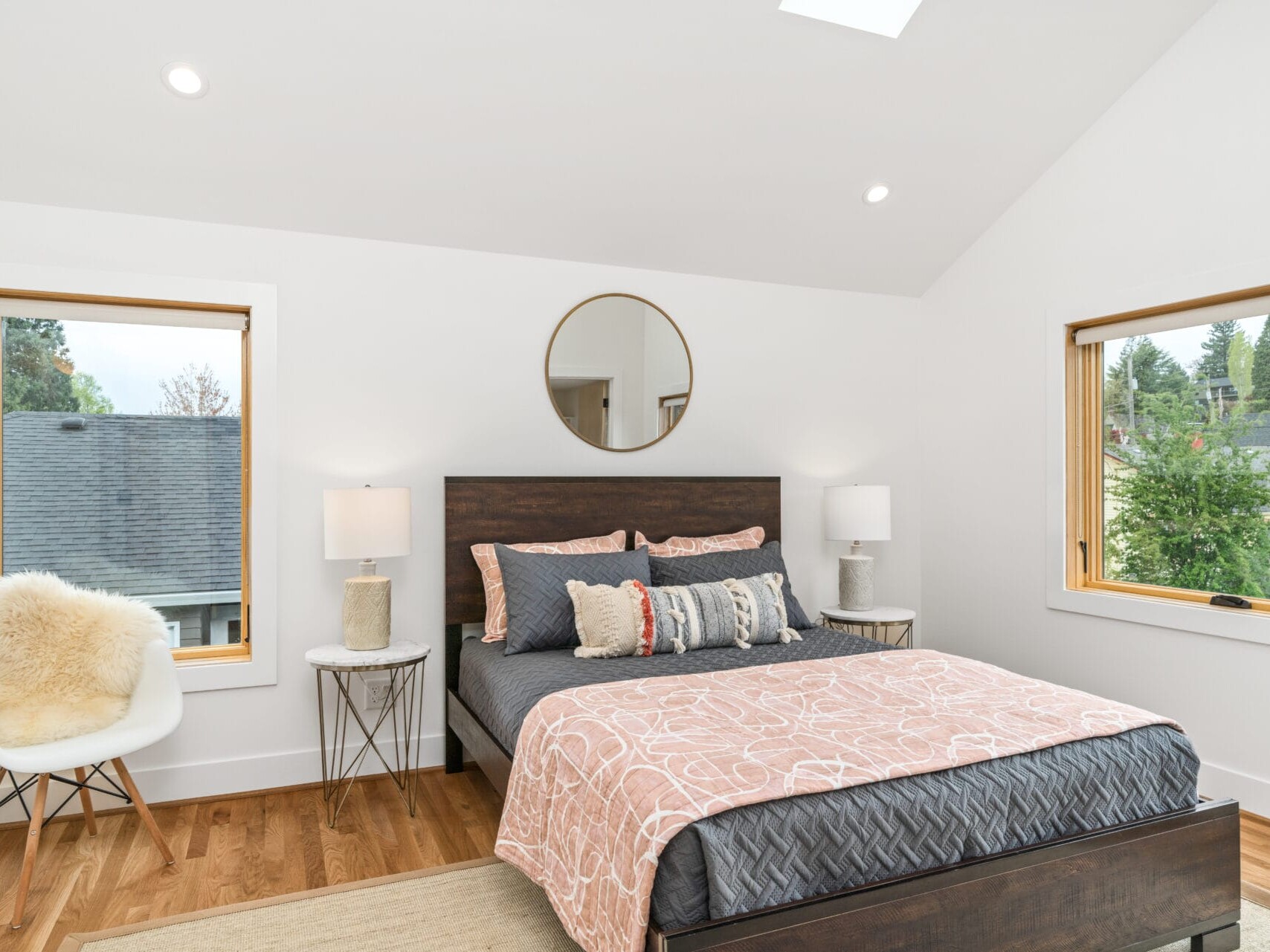 A bright Portland, Oregon bedroom features a double bed with pink patterned bedding and a dark wood headboard. Two side tables with lamps flank the bed, while a round mirror adds charm. A white furry chair by the window complements the wooden flooring as natural light pours in through two large windows.