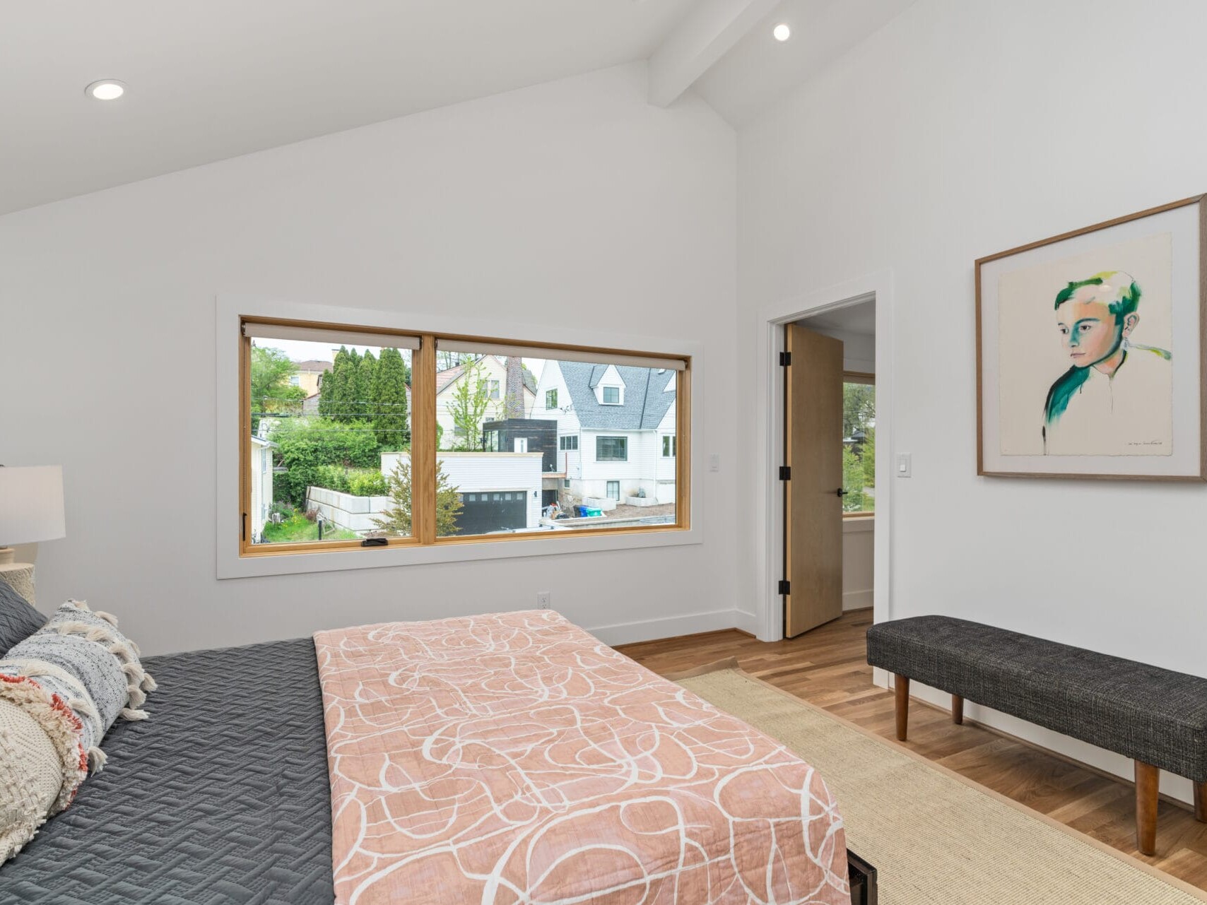 A minimalist bedroom in Portland, Oregon, features a large window with views of houses and greenery. The room includes a bed with patterned linens, a bench, a colorful portrait on the wall, and wooden flooring, all creating a bright and airy atmosphere.