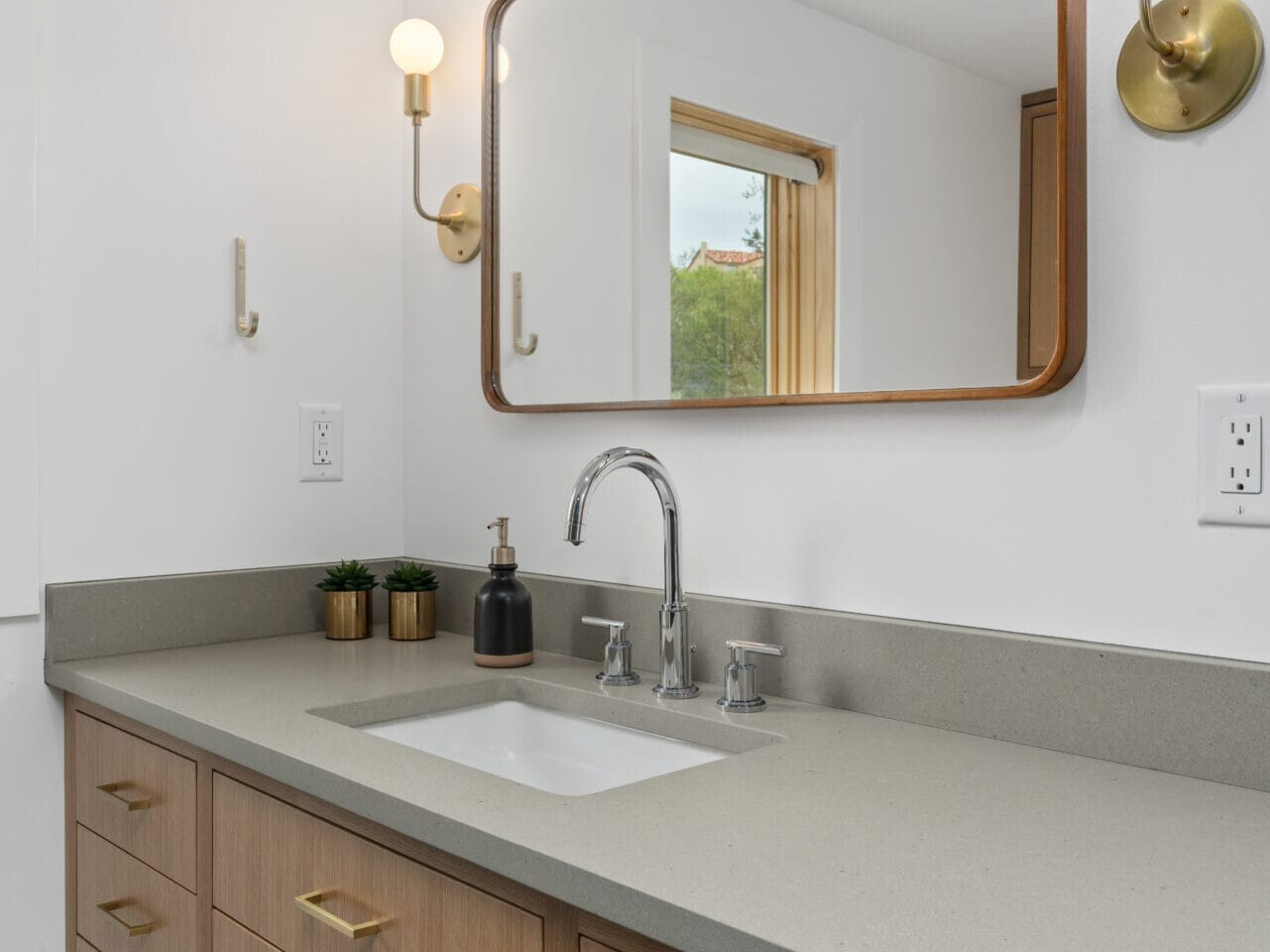 In a modern Portland, Oregon bathroom, a large rectangular mirror with a wooden frame hangs above a gray countertop with a white sink. Brass faucets and cabinet handles match the wall-mounted light fixtures on either side. Two small plants sit near the sink, adding a touch of nature.