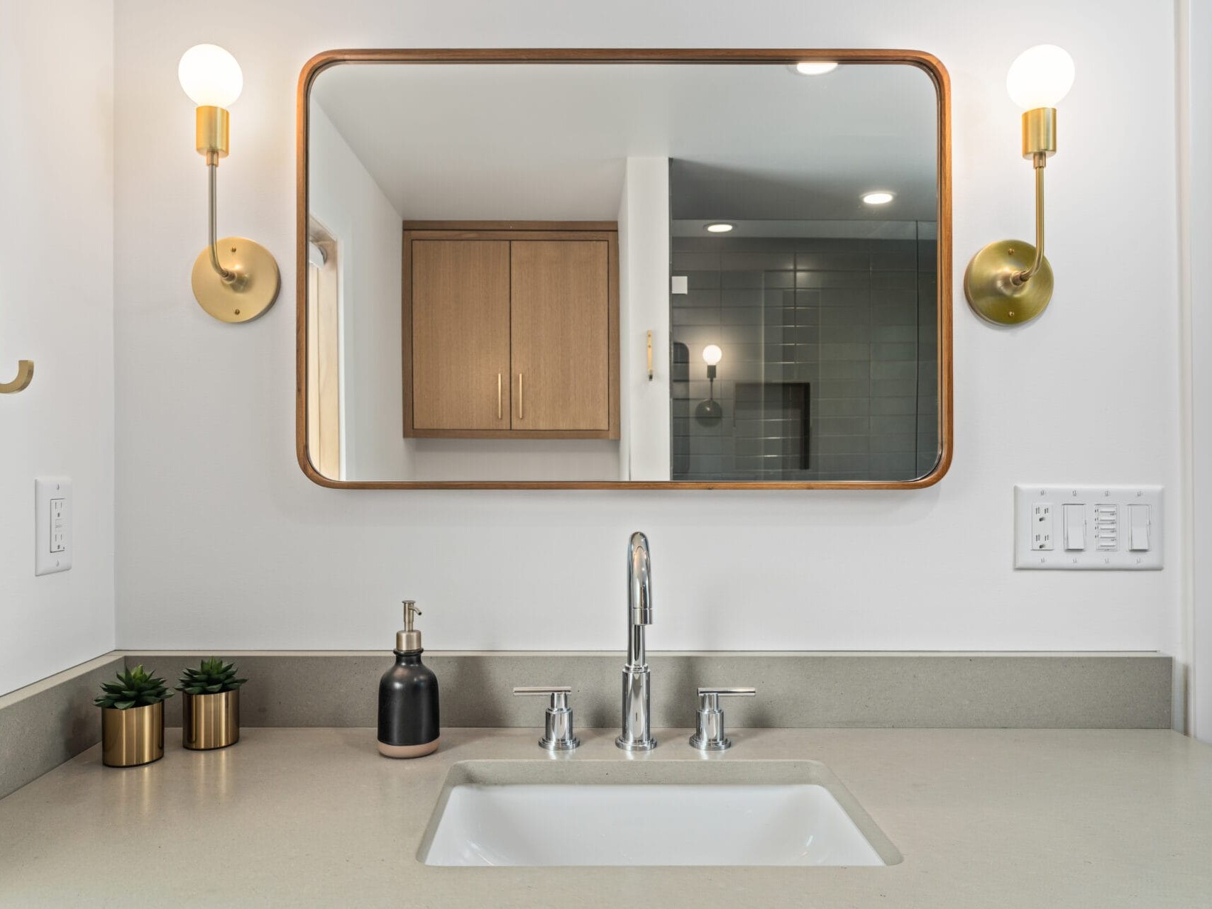 A modern bathroom vanity in Portland, Oregon features a rectangular mirror with brass sconces on either side, a sleek faucet, and a beige countertop with a built-in sink. Two small potted plants and a soap dispenser are placed on the counter.