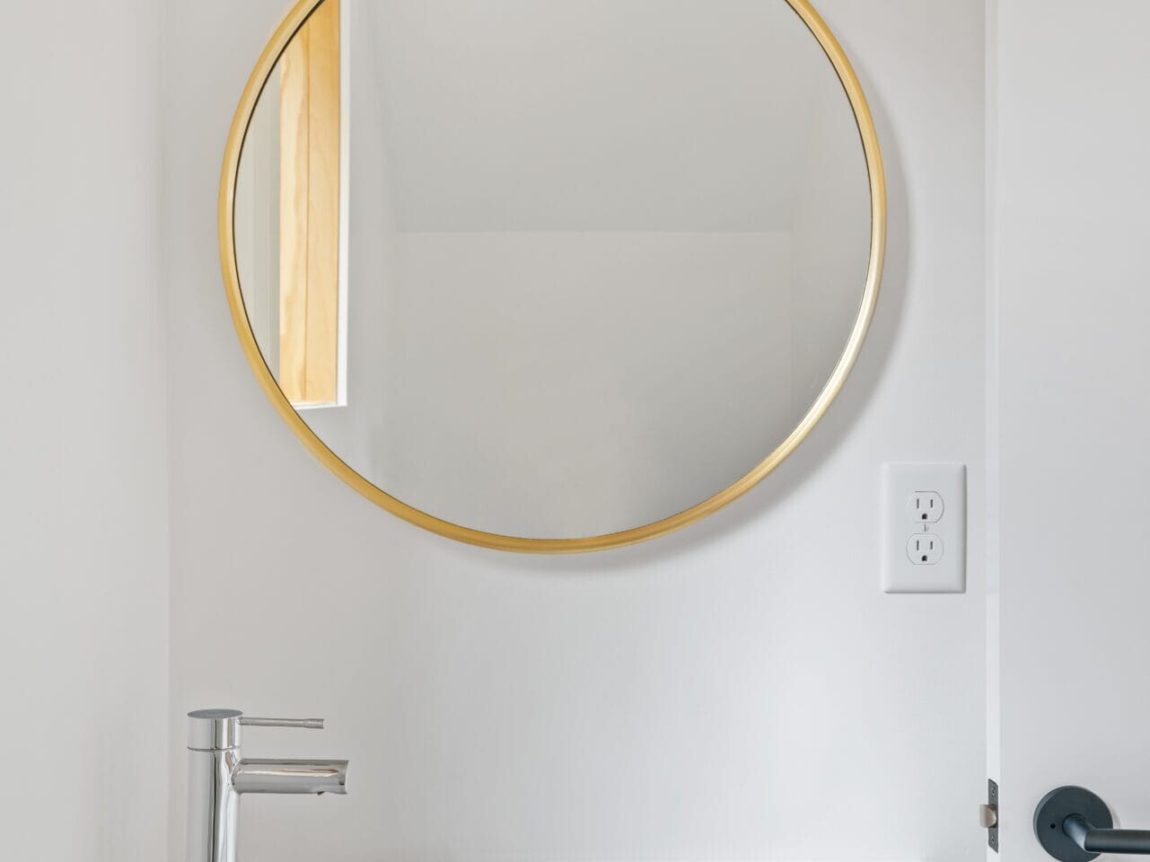 A minimalist Portland, Oregon bathroom features a white rectangular sink with a sleek silver faucet. Above the sink, a round mirror with a gold frame is mounted on a white wall. A door is slightly ajar on the right side of the image.
