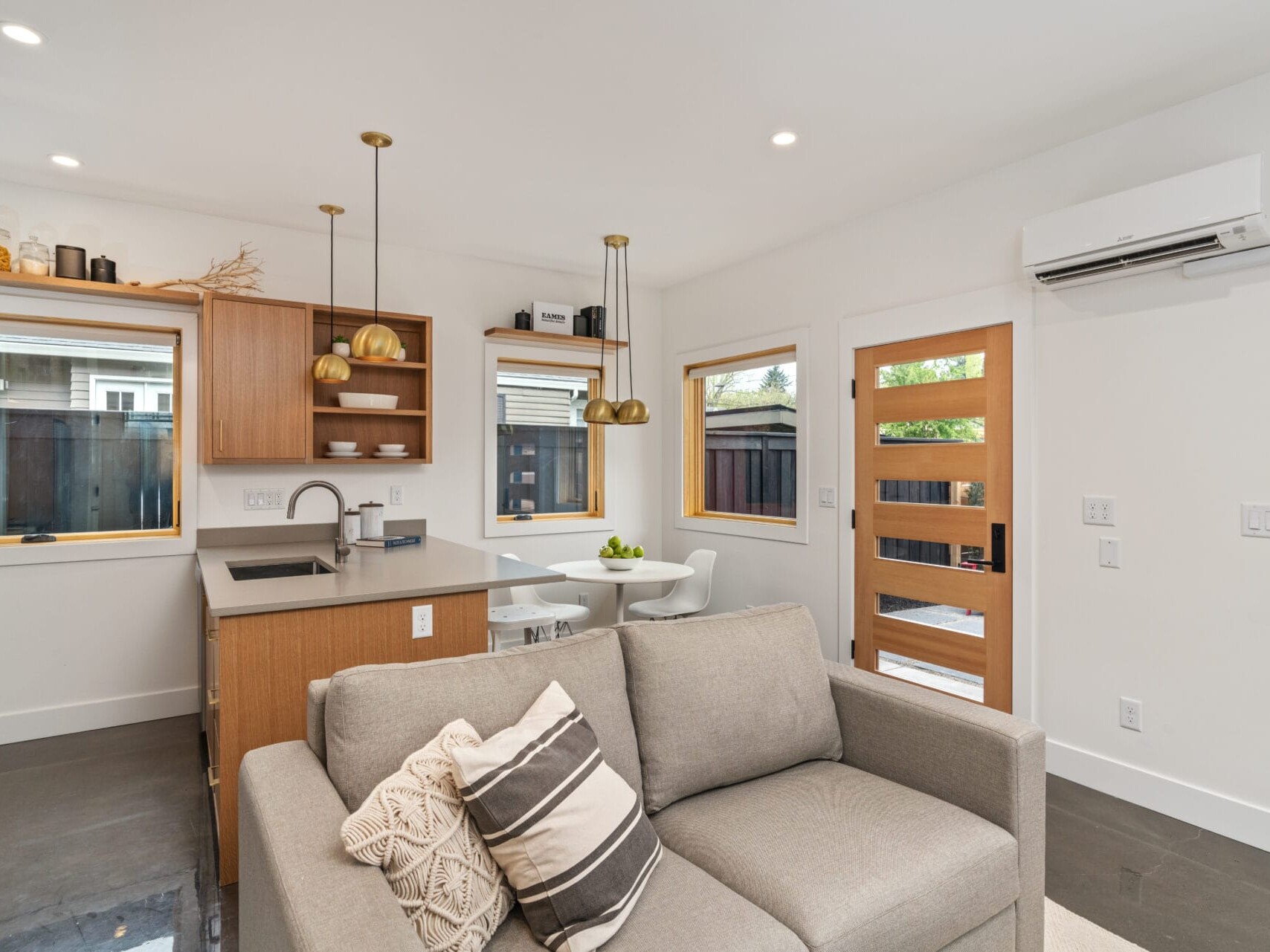 A modern open-plan living space in Portland, Oregon features a gray sofa with patterned cushions, a kitchen with wooden cabinetry, pendant lights, a dining area with a round table, and large windows. A wooden front door and a white wall-mounted air conditioner complete the serene ambiance.