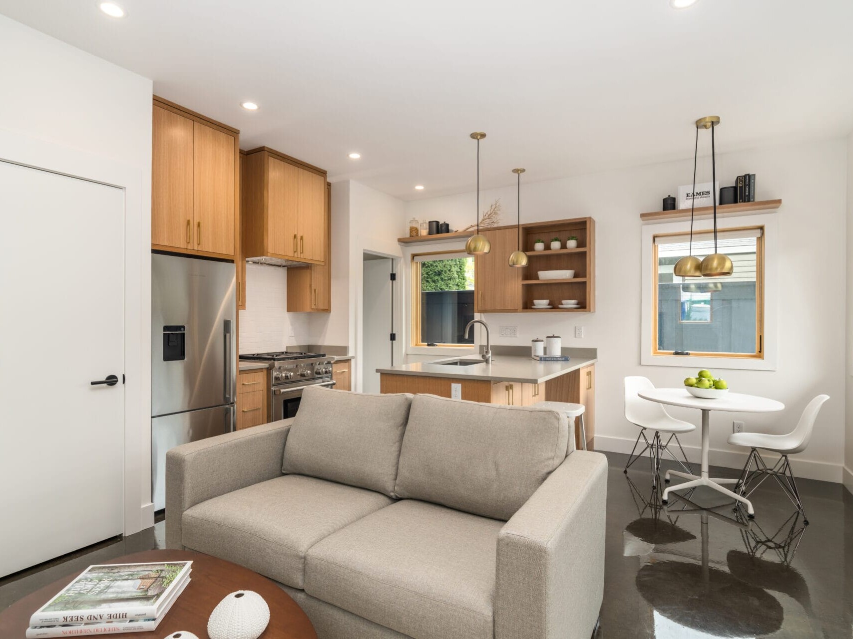 Modern kitchen and living area in Portland, Oregon, with natural light. Features a gray sofa, wooden cabinets, stainless steel appliances, and a small dining table with white chairs. Pendant lights hang above the kitchen island. Large windows brighten the space.
