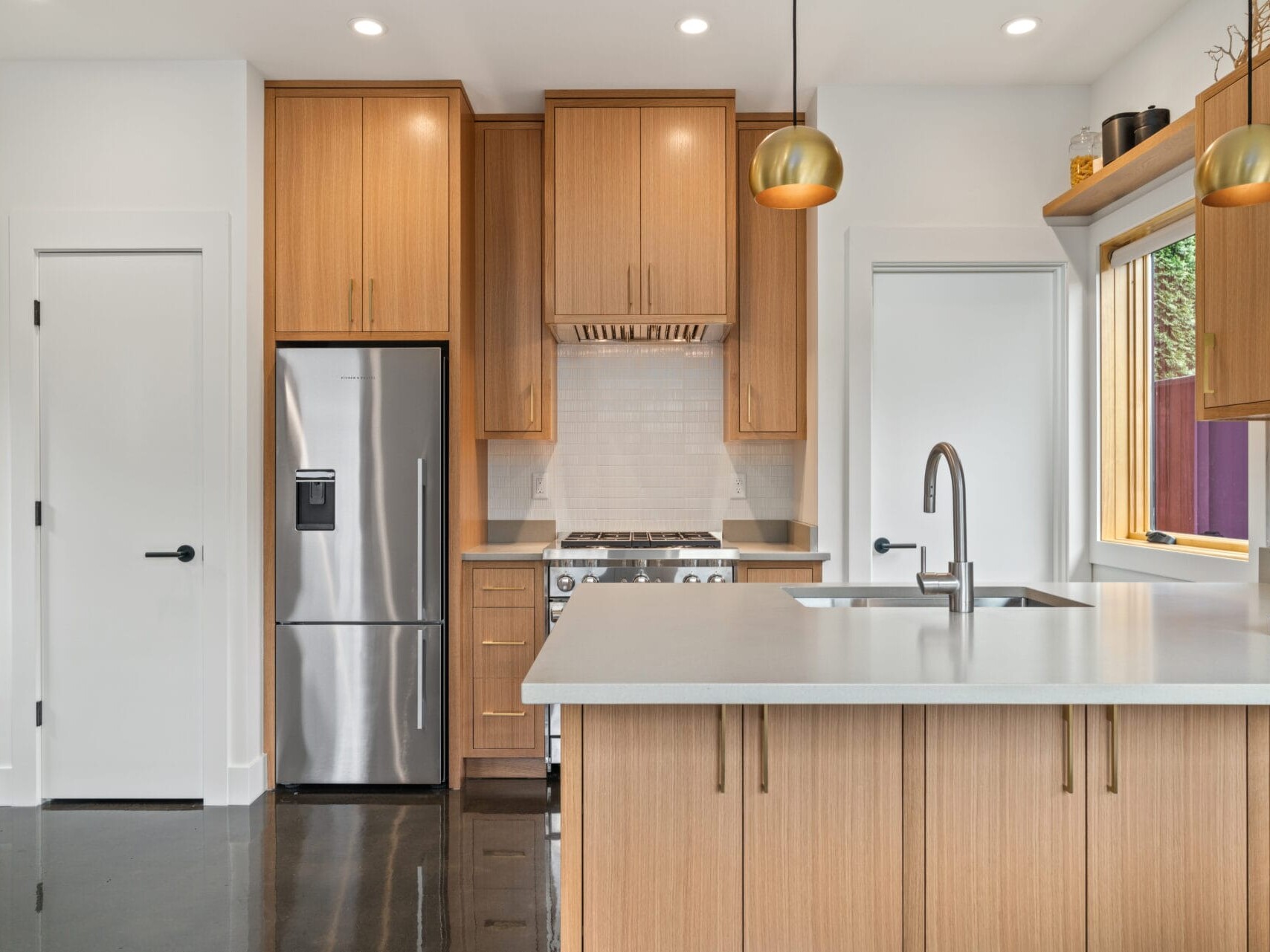 Modern kitchen in Portland, Oregon, featuring wooden cabinets, a stainless steel fridge, and a built-in oven. A white countertop island with a sink is centered beneath two hanging pendant lights. A window above the sink floods the space with natural light.