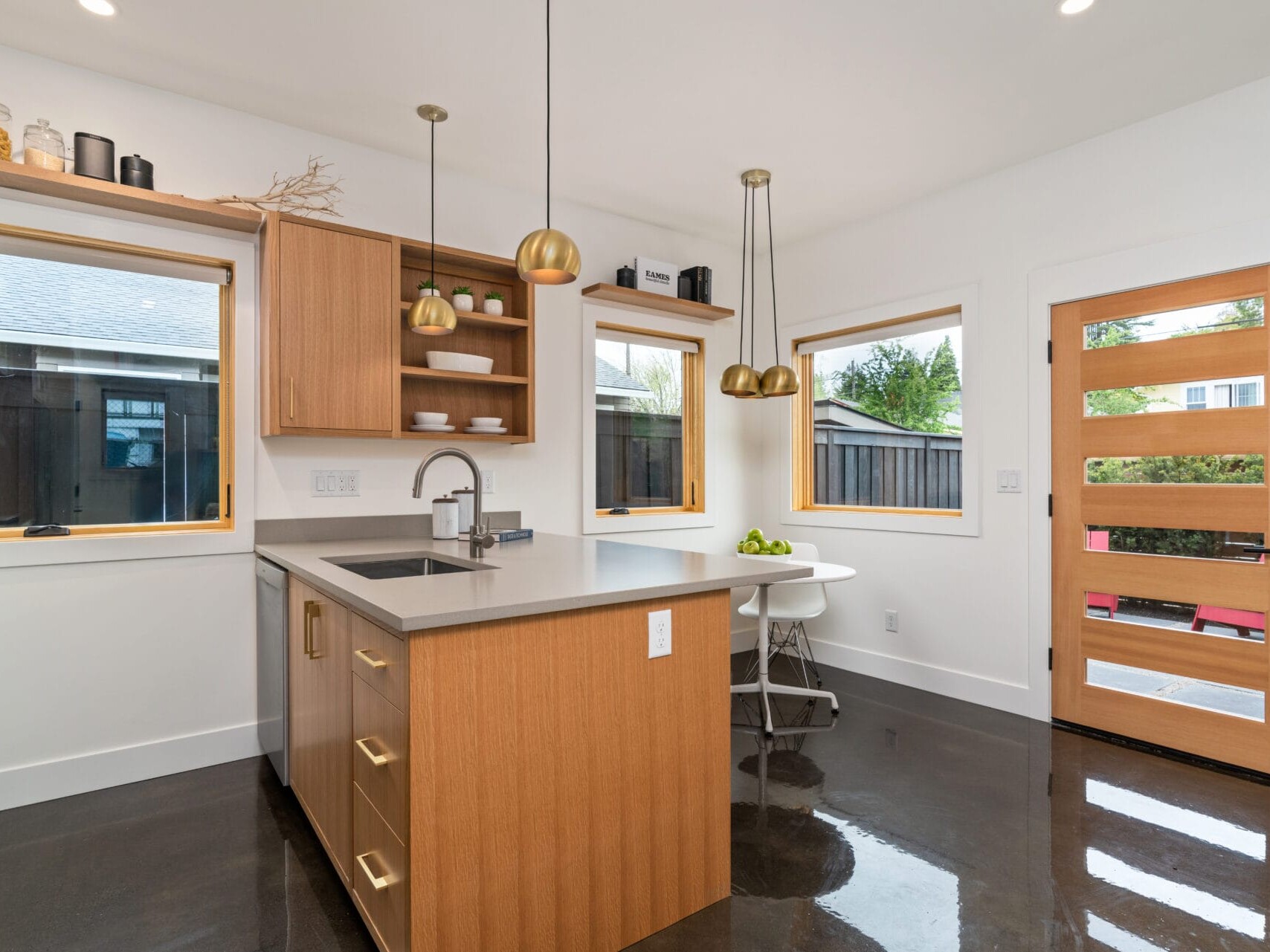 A modern kitchen in Portland, Oregon, features a central island with wooden cabinets and a gray countertop. Three pendant lights hang above the island, complemented by windows with wooden frames and a table for two near a wooden door with horizontal slats.