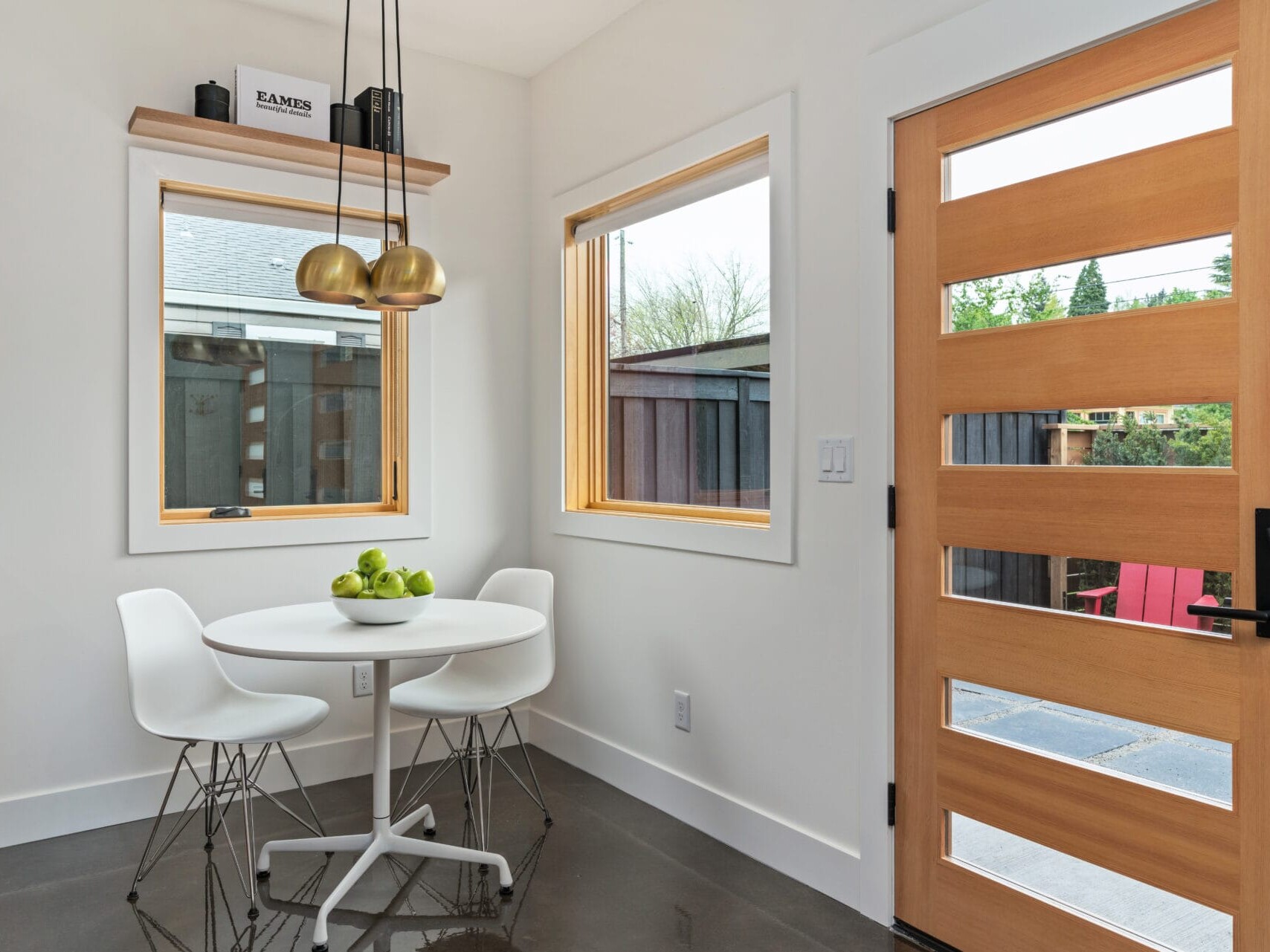 A modern dining area in Portland, Oregon, features a white round table with four chairs on a sleek dark floor. Green apples sit invitingly on the table. Two large windows and a wooden front door with glass panels add charm, while a set of hanging lights above completes the decor.