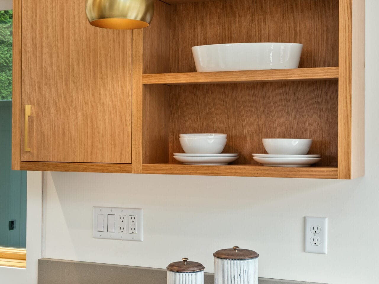 A modern kitchen in Portland, Oregon features wooden cabinets and open shelving displaying white bowls and small potted plants. A brass pendant light hangs above. The countertop holds two ceramic containers and a cookbook titled Taste & Technique.
