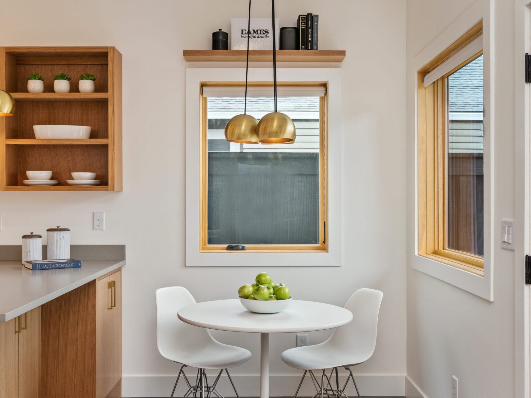 A modern kitchen nook in Portland, Oregon features a white round table and two matching chairs. A bowl of green apples sits invitingly on the table. To the left, wooden cabinets with open shelving add warmth, while pendant lights illuminate the space through a sunlit window.
