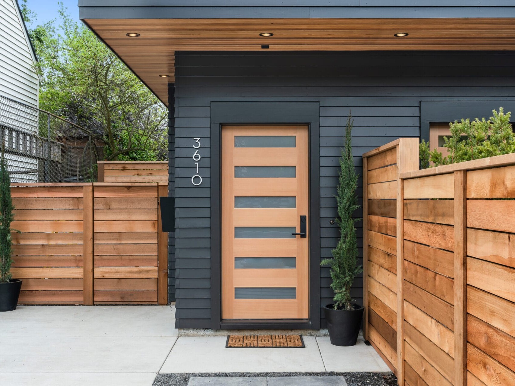 The modern house entrance in Portland, Oregon, boasts a wooden door with frosted glass panels, framed by elegant horizontal wooden fencing. Two tall potted plants gracefully flank the entrance, while the house number 3610 stands out on the sleek dark gray exterior wall.