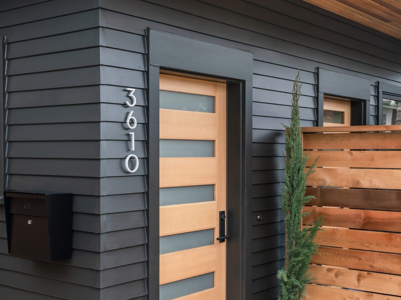 A modern house entrance in Portland, Oregon features a black and wood-themed door, numbered 3610. The exterior boasts horizontal black paneling and a wooden overhang. A potted plant is nestled near a wooden privacy screen beside the door, adding a touch of nature to this contemporary facade.