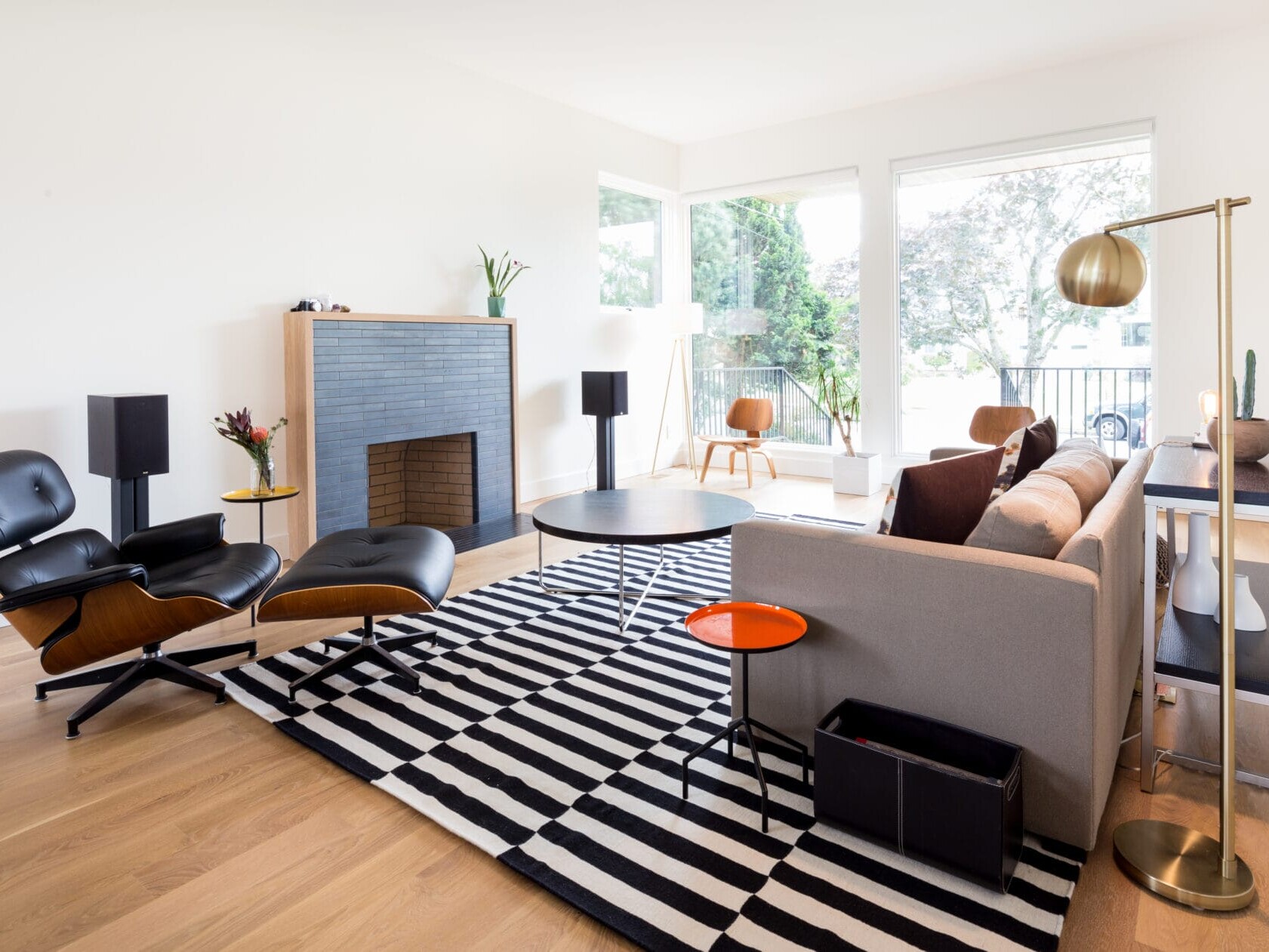 A modern living room in Portland, Oregon features a black and white striped rug, a mid-century chair with a matching ottoman, a gray sofa, a round coffee table, and a small fireplace. Large windows invite natural light to fill this cozy Northwest space.
