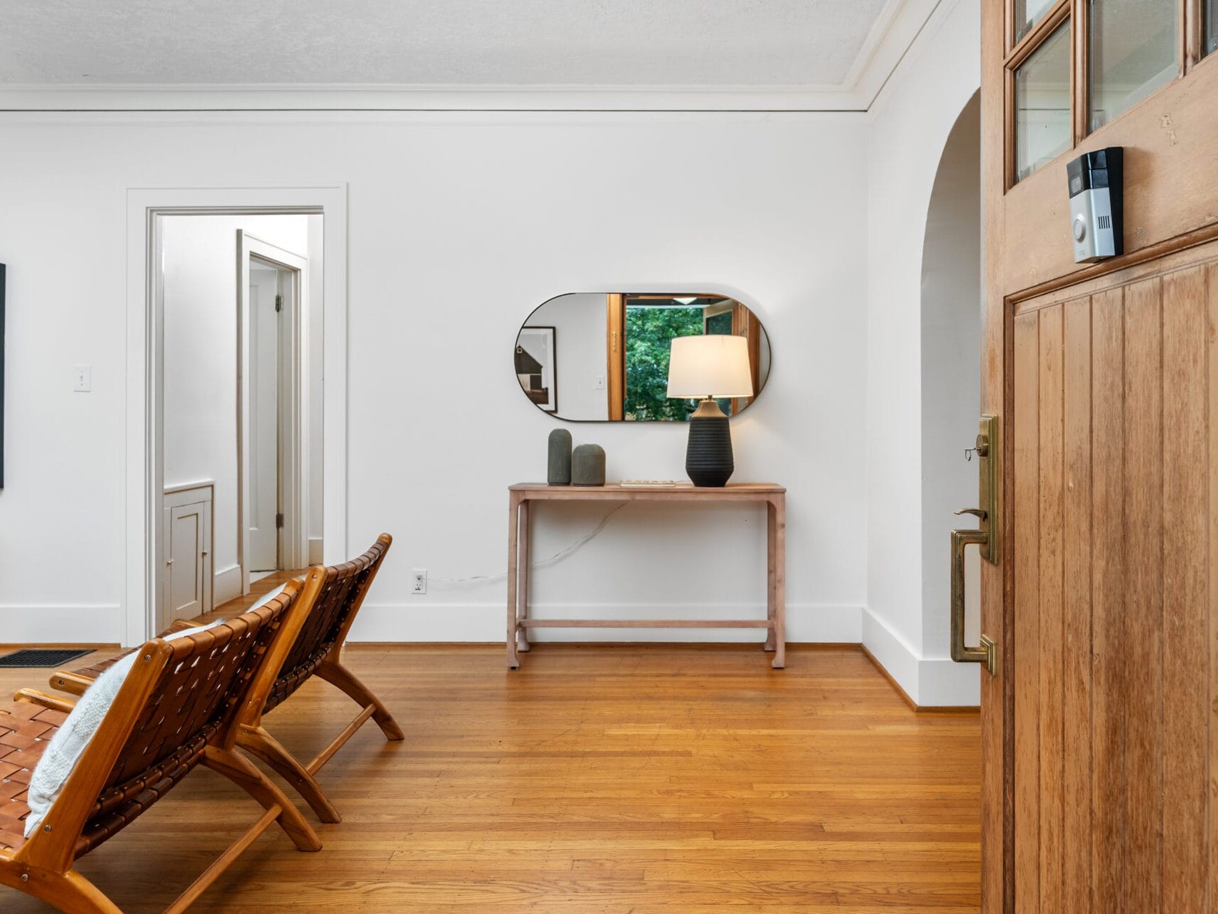 A cozy, minimalist living room with wooden flooring. Two woven chairs face a wooden console table against a white wall. An oval mirror and a lamp sit on the table. An open doorway and a partially visible wooden door are also present.