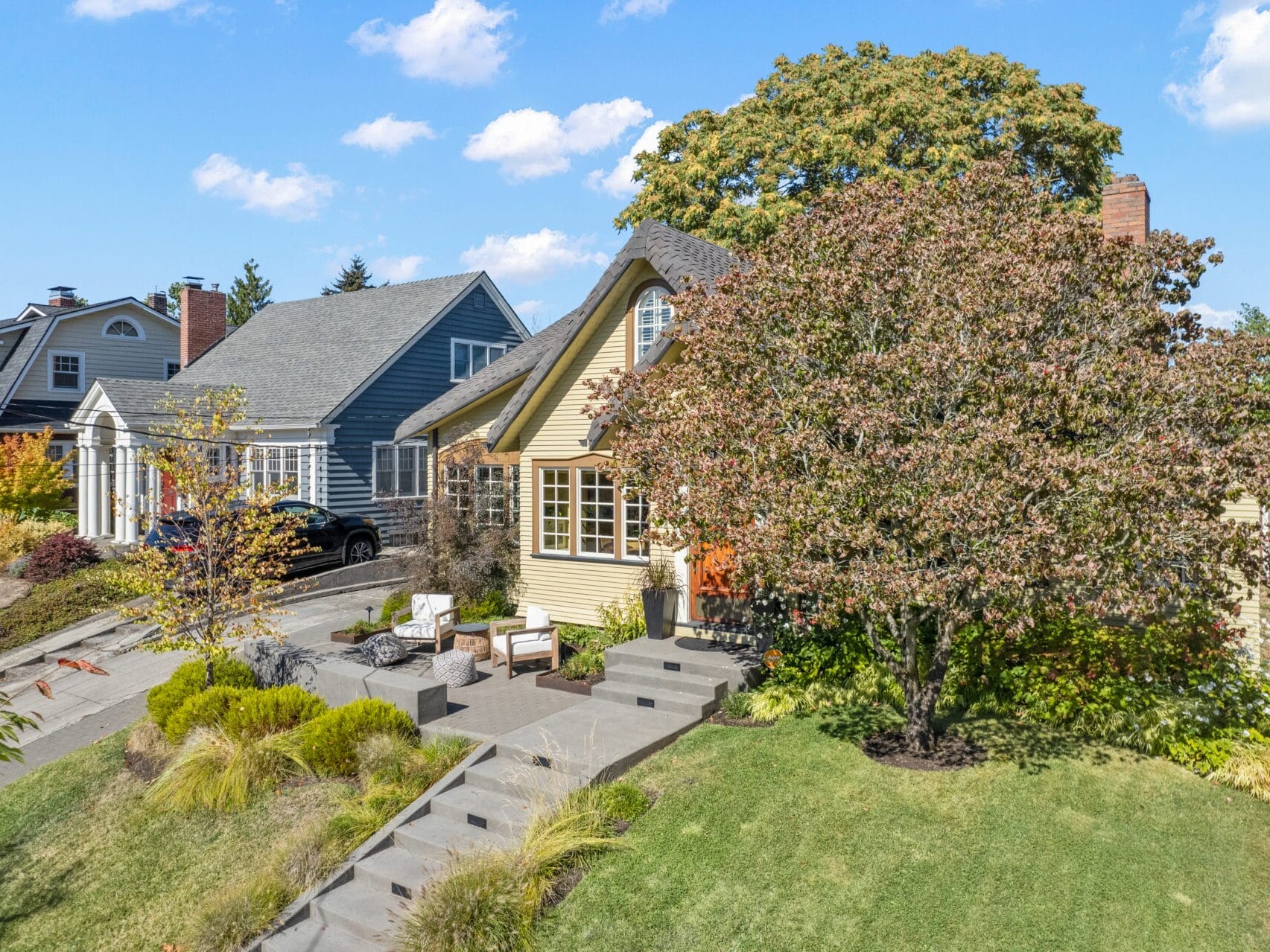 A charming suburban home with yellow siding, surrounded by greenery. A well-maintained front yard features a tree and various shrubs. Outdoor furniture sits on the front porch under a clear blue sky. Other houses are visible in the background.