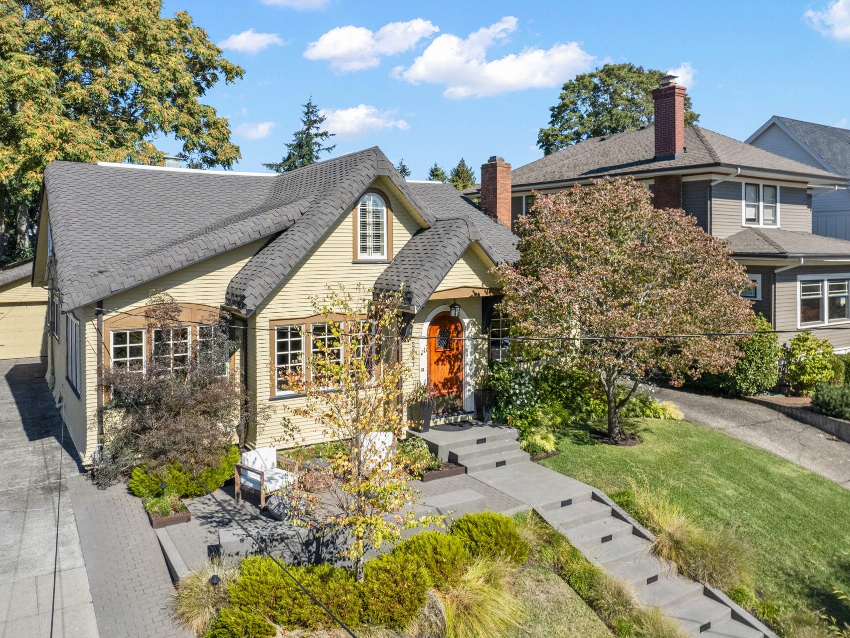 A charming two-story house with beige siding, a gray roof, and an orange front door sits in a suburban neighborhood. It has a manicured lawn, a paved driveway leading to a detached garage, and is surrounded by trees and shrubs under a blue sky.