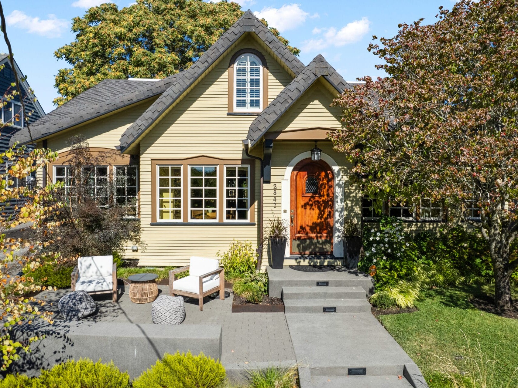 A charming, light yellow cottage-style house with a gray shingled roof and arched doorway. A stone walkway leads through a landscaped front yard with trees and shrubs, and two white chairs sit by the entrance. Sunny day with a blue sky.