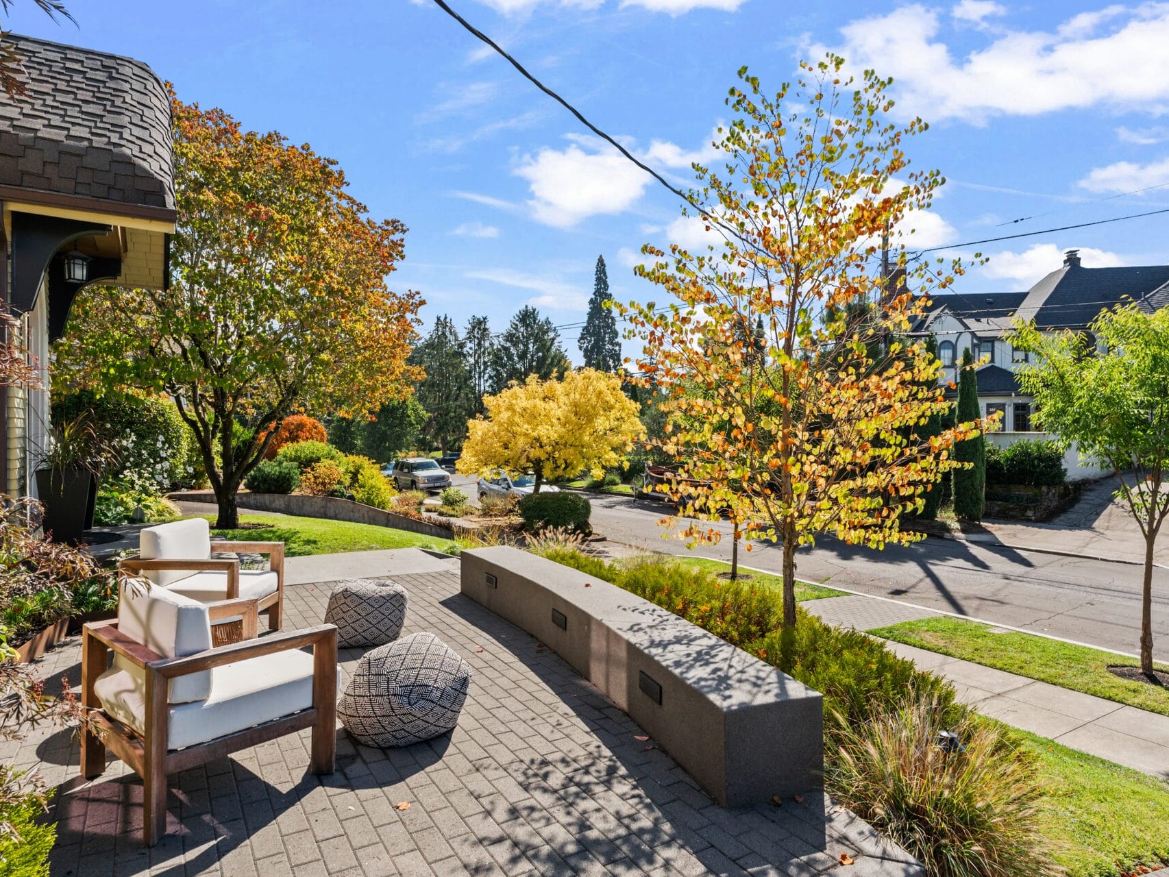 A sidewalk patio with two chairs and a bench overlooks a suburban street lined with colorful autumn trees. The sky is clear and blue, and houses are visible in the background. The scene is bright and inviting.