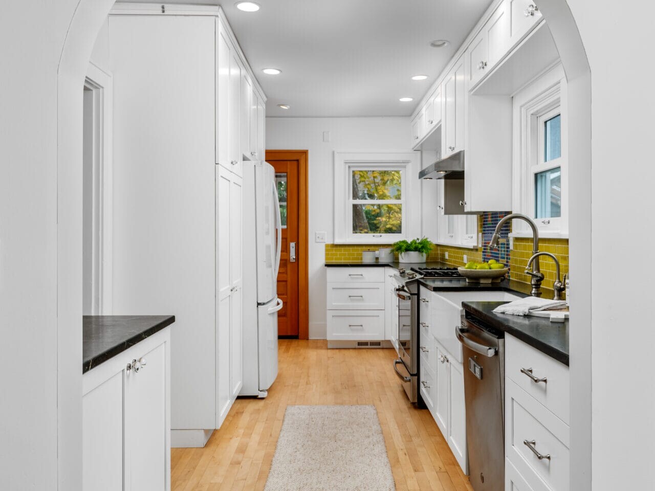 A modern kitchen with white cabinetry, black countertops, and stainless steel appliances. A long tan rug runs along the wooden floor. The backsplash is yellow-green, and a window with a view of greenery is centered above the sink.