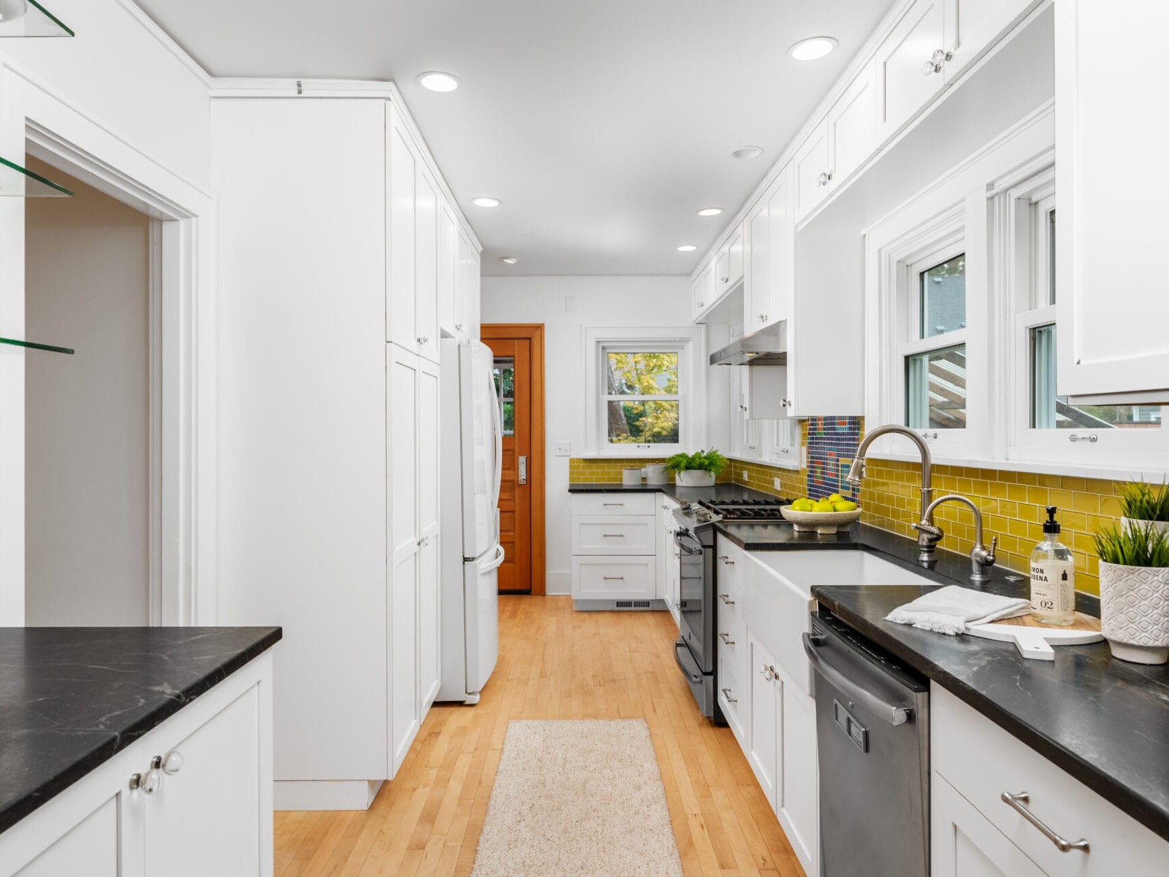A modern kitchen with white cabinets, black countertops, and a wooden floor. Yellow tile backsplash adds a pop of color. Appliances include a refrigerator, dishwasher, and oven. A window provides natural light, and potted plants add greenery.