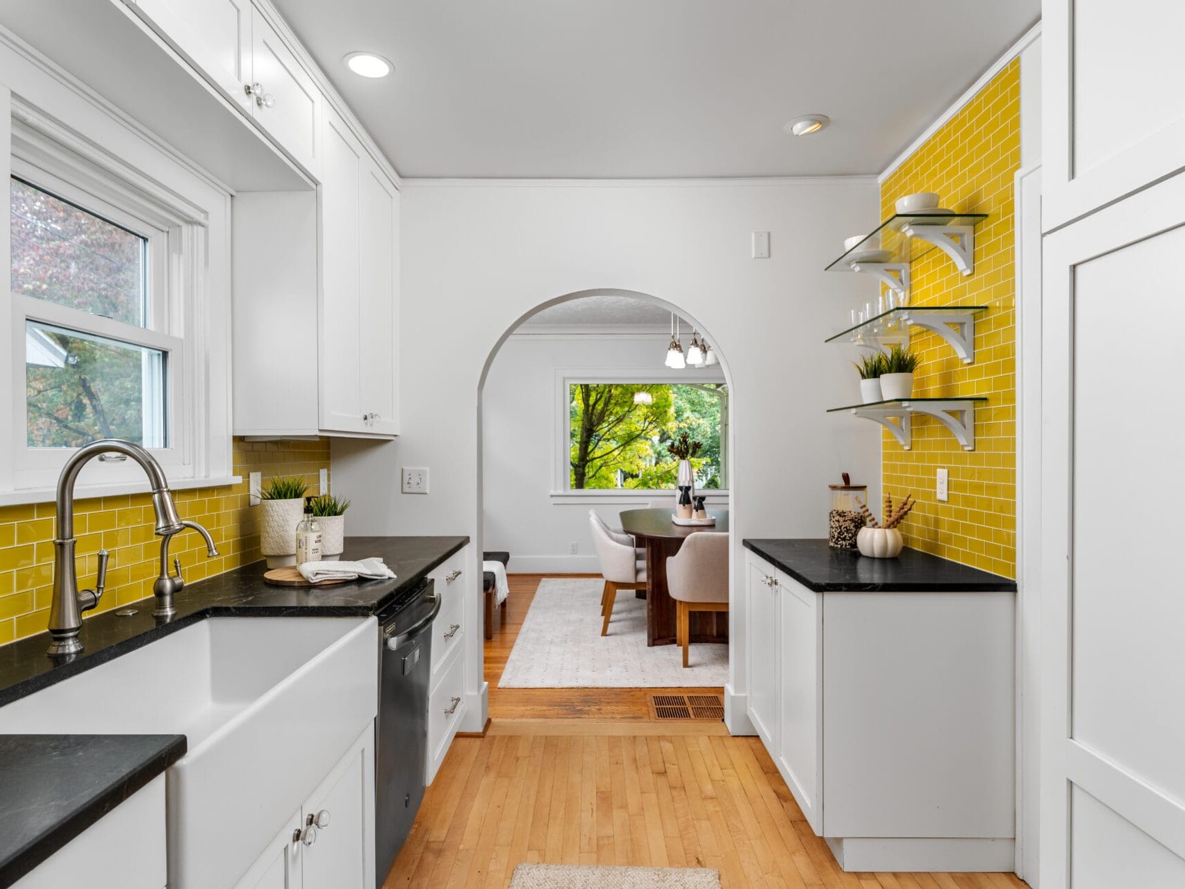 A modern kitchen with white cabinets and black countertops features a yellow tile backsplash. An arched doorway leads to a dining area. Wooden floors and green plants add warmth to the bright, open space.