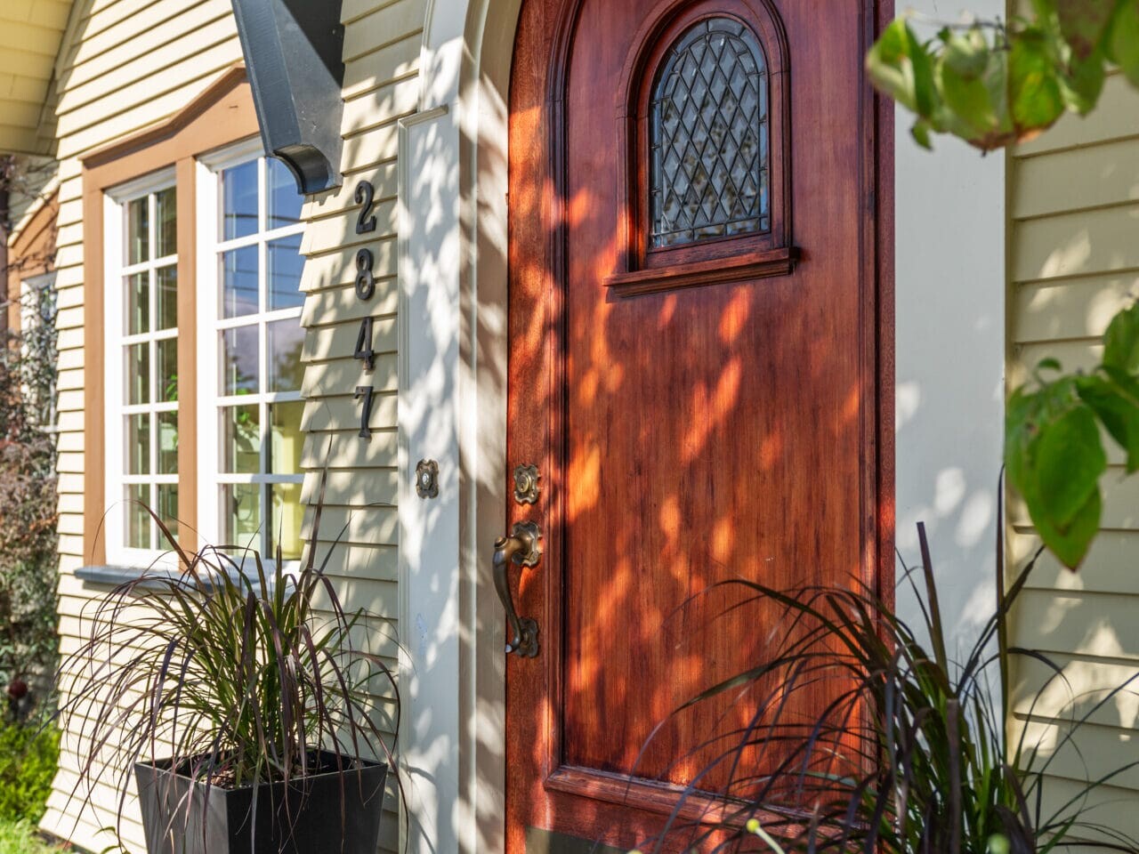 A charming arched wooden door with a small window is framed by two planters on a porch. Sunlight filters through nearby leaves, casting shadows on the pale yellow exterior walls. The door has an ornate handle and a lantern above.