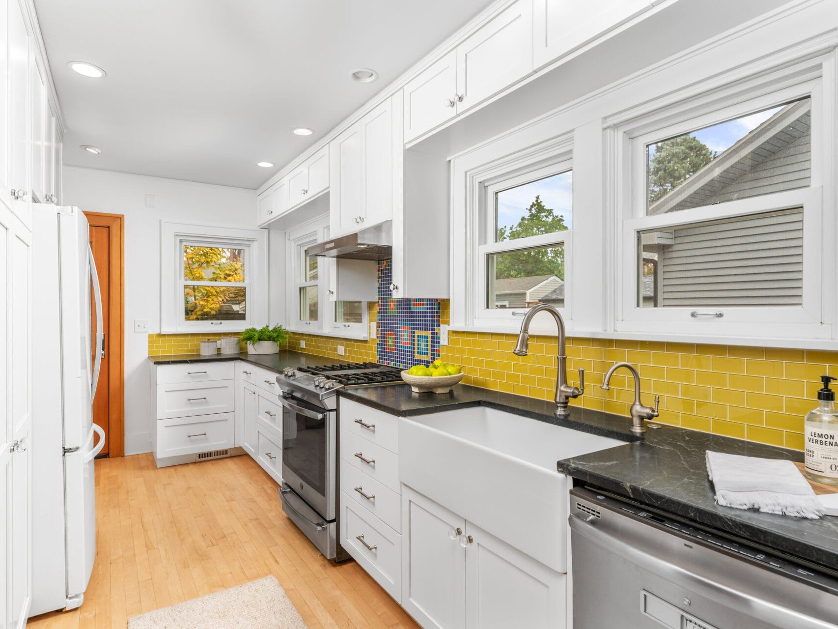 A modern kitchen with white cabinets, black countertops, and yellow tile backsplash. There are stainless steel appliances, a bowl of bananas, and a Thank you hand soap by the sink. Windows provide natural light.