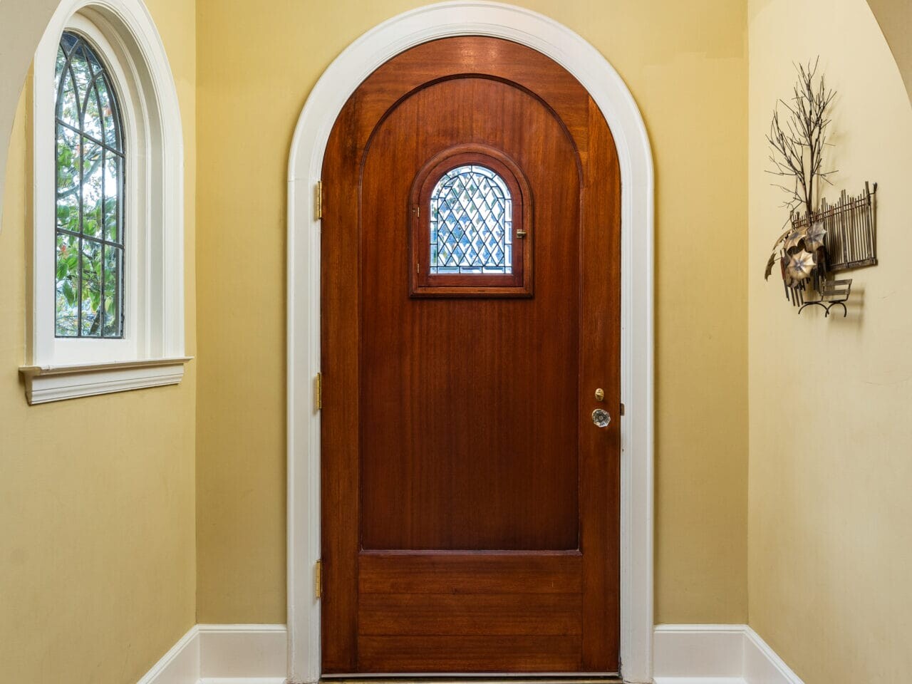 A wooden arched front door with a small window in the center, set in a cozy entryway with a light yellow wall. A window with an arched top is on the left, and a woven rug lies on the wooden floor beneath. A light fixture hangs from the ceiling.