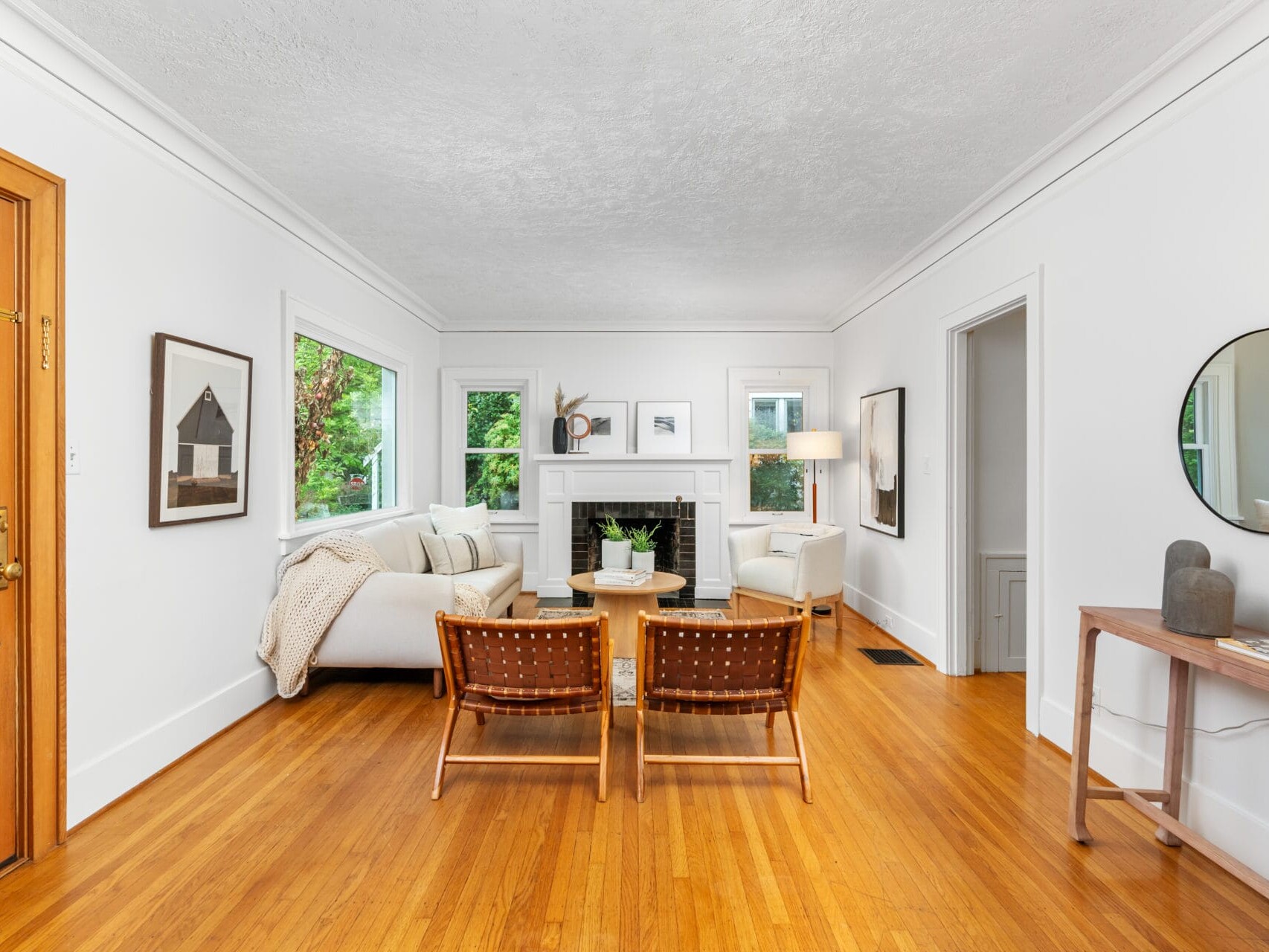 A cozy living room with white walls and a wooden floor. It features a white sofa, two brown woven chairs, a round coffee table, and a fireplace. Art adorns the walls, and three windows bring in natural light. A wooden console holds a round mirror.