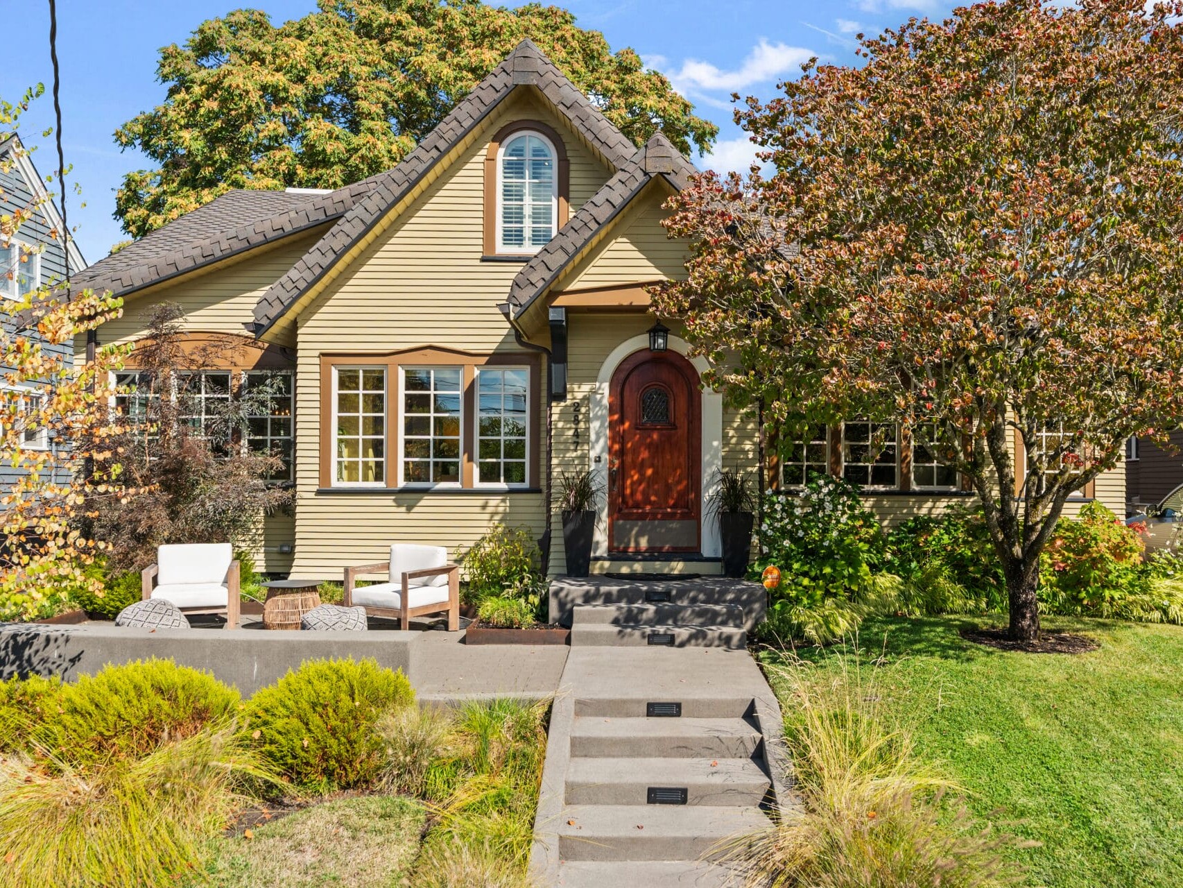 Charming cottage-style house with a steeply pitched roof and a red front door, surrounded by a well-manicured garden. Two white chairs on the front patio and a tree with autumn foliage enhance the inviting look. Steps lead up from a lush green lawn.
