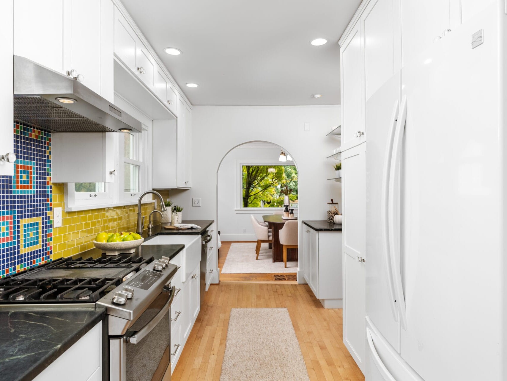 A modern kitchen with white cabinets, a black countertop, and colorful mosaic backsplash. It features a stainless steel stove and a bowl of lemons. The space opens into a dining area with wooden floors and a view of greenery through a window.