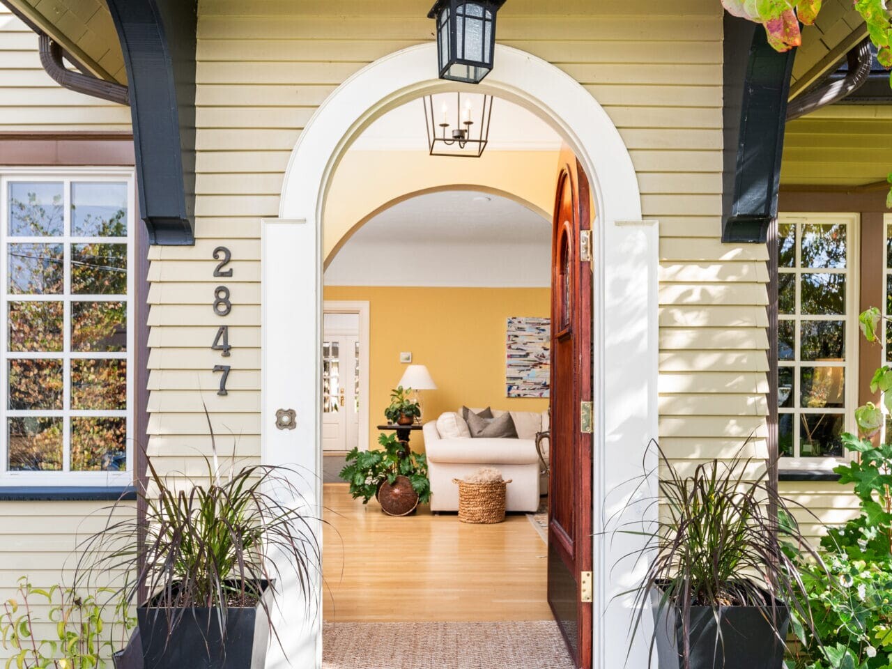 A welcoming entrance to a home with a yellow exterior. Theres an arched wooden door, black sconces, and potted plants. Inside, a bright, cozy living room with a plush white sofa is visible through the entrance.