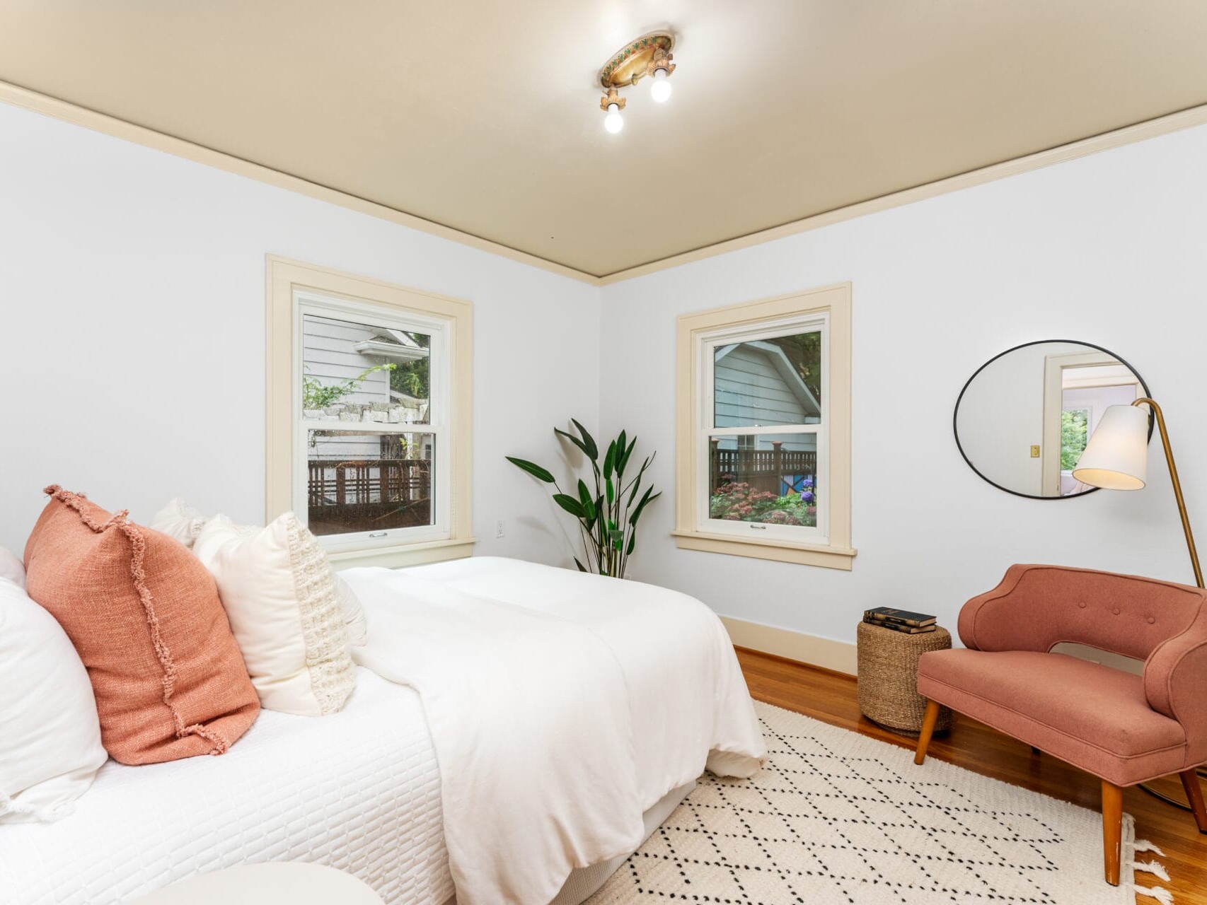 A cozy bedroom featuring a white bed with pink and white pillows. A pink armchair sits beside a round mirror and a tall floor lamp. There are two windows with outdoor views, a potted plant, and a light fixture on the ceiling.